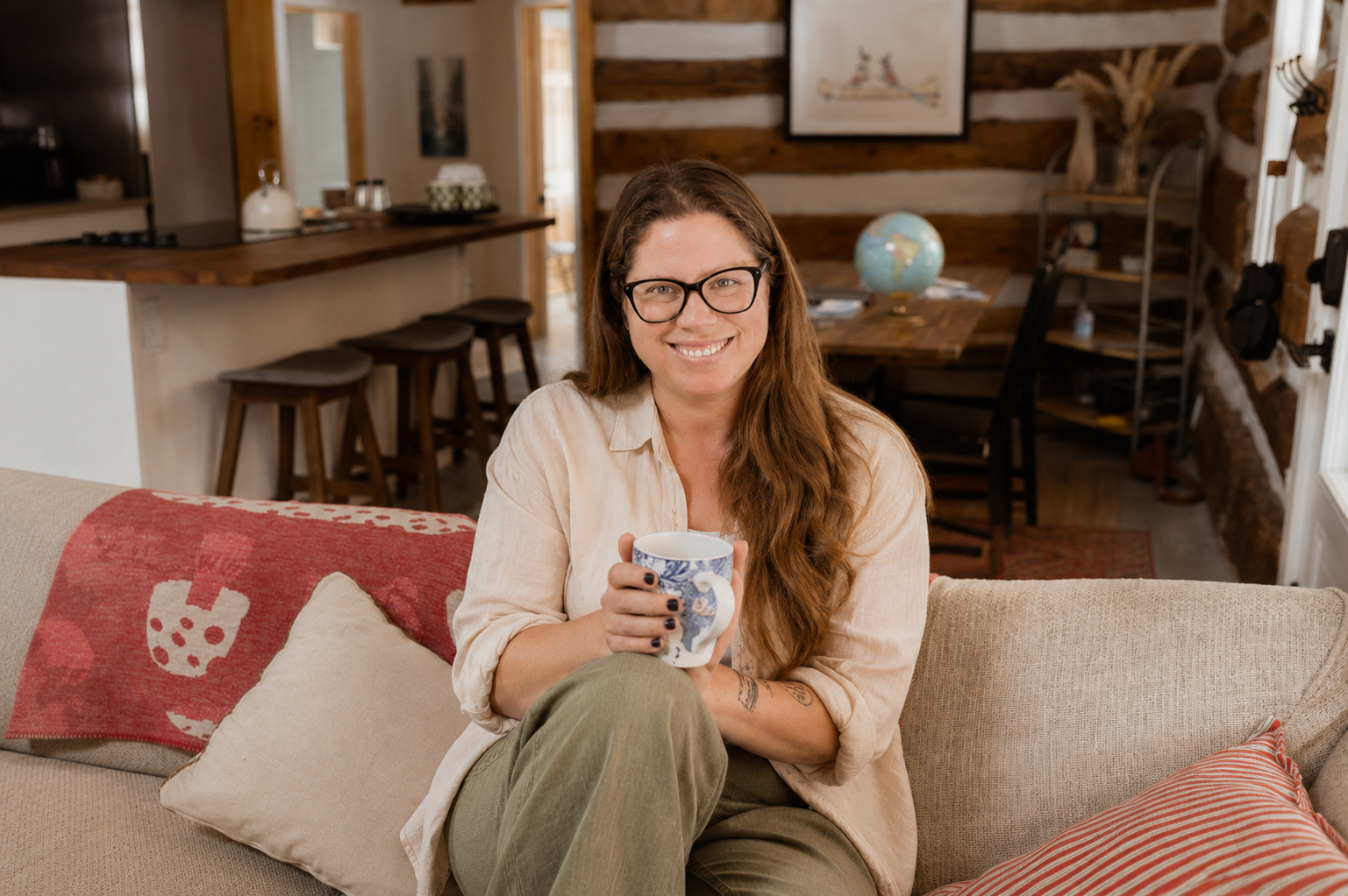 A woman with long light brown hair, glasses, and a beige shirt smiling and holding a mug, sitting on a beige couch in a cozy, rustic home interior.
