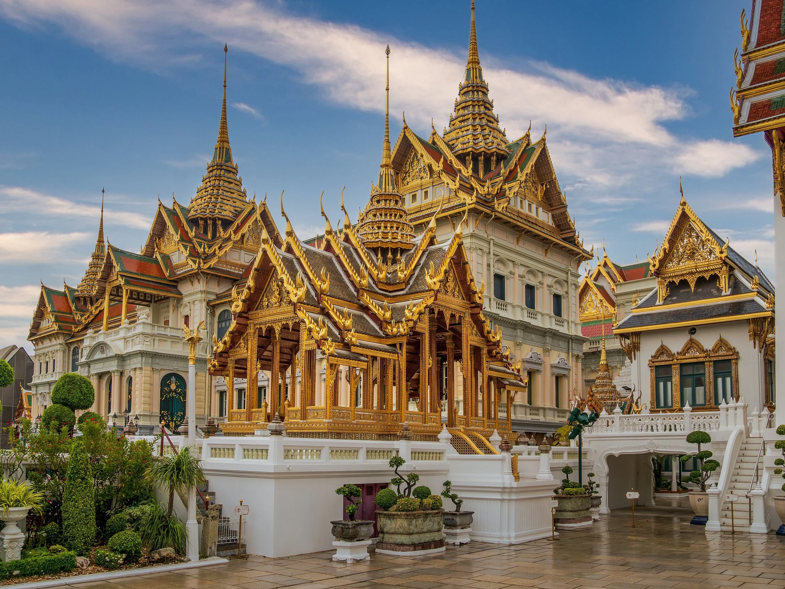 A grand Thai-style palace with ornate golden rooftops and spires, surrounded by manicured greenery and a stone-paved courtyard under a partly cloudy sky.