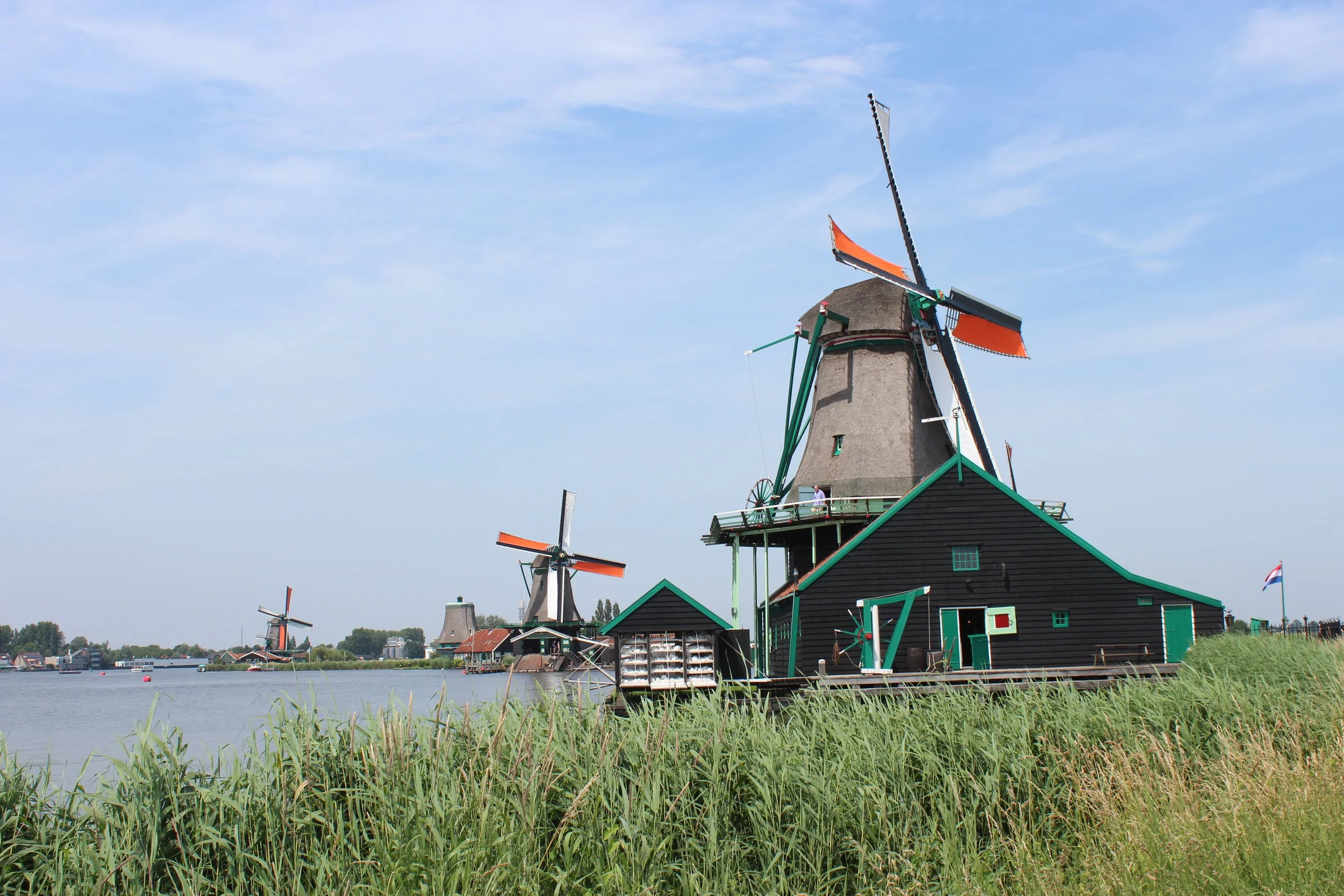 A traditional Dutch windmill with orange and black blades near a river, with other windmills and buildings visible in the background and grassy foreground.