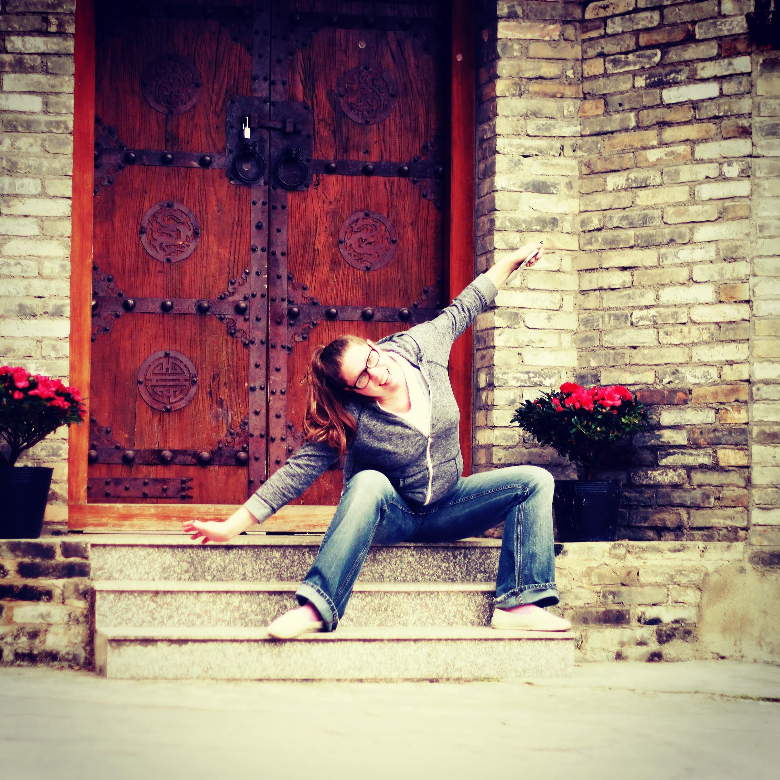 A woman with long red hair, glasses, and a gray sweater, lying on her side on brick stairs in front of a large wooden door with metal accents, surrounded by brick walls and potted plants with red flowers.