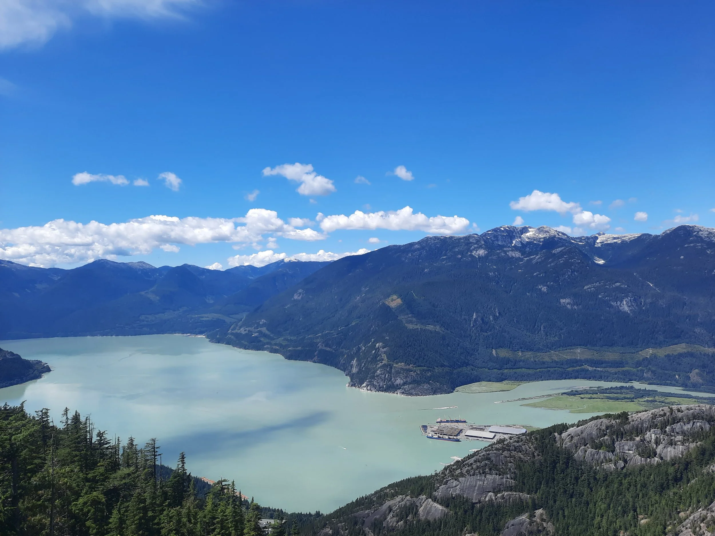 A large body of light blue-green water in a mountainous landscape with green forests, rugged mountains with some snow on the peaks, and a partly cloudy sky overhead. There is a ship docked near a dock in the foreground in Squamish British Columbia