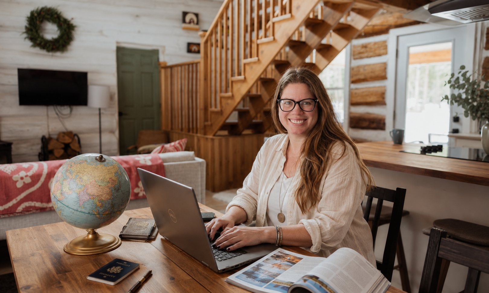 A woman with long light brown hair and glasses sitting at a wooden table in a cozy interior, using a laptop with an open magazine, a notebook, and a globe on the table, smiling at the camera.