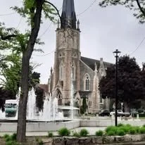 A historic church with a tall steeple, surrounded by trees and a fountain in front.