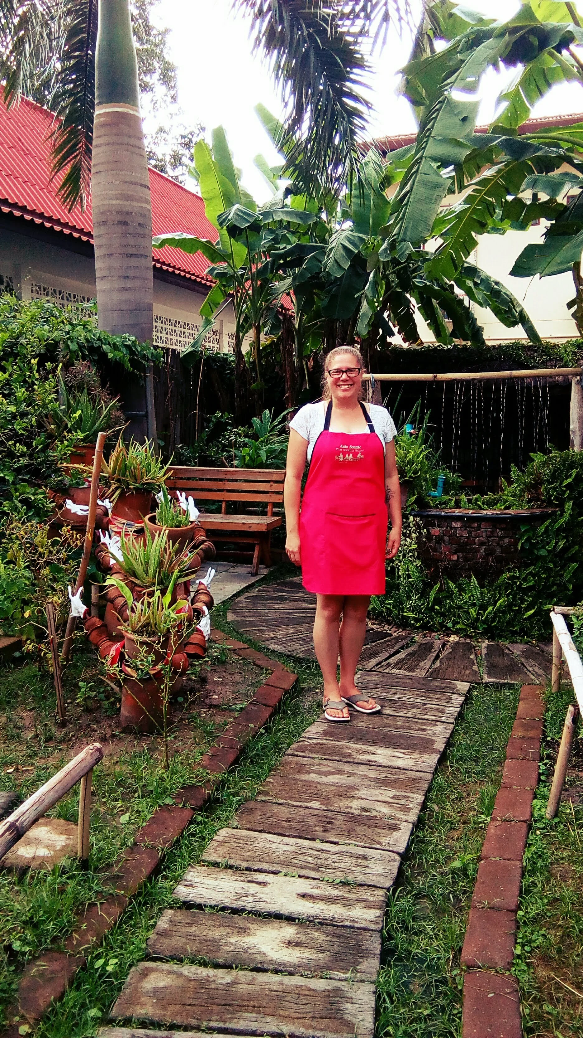 A smiling woman wearing glasses, a white t-shirt, and a pink apron, stands on a wooden pathway in a lush garden with large green plants, potted succulents, a bench, and red roof house in the background, outside a cooking class in Chiang Mai Thailand