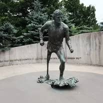 Bronze statue of a Terry Fox running, mounted on a small base with foliage, outdoors with trees in the background at mile 1 marker in St Johns Newfoundland.