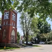 A red brick church with a tall bell tower, surrounded by green trees and a sidewalk in front.