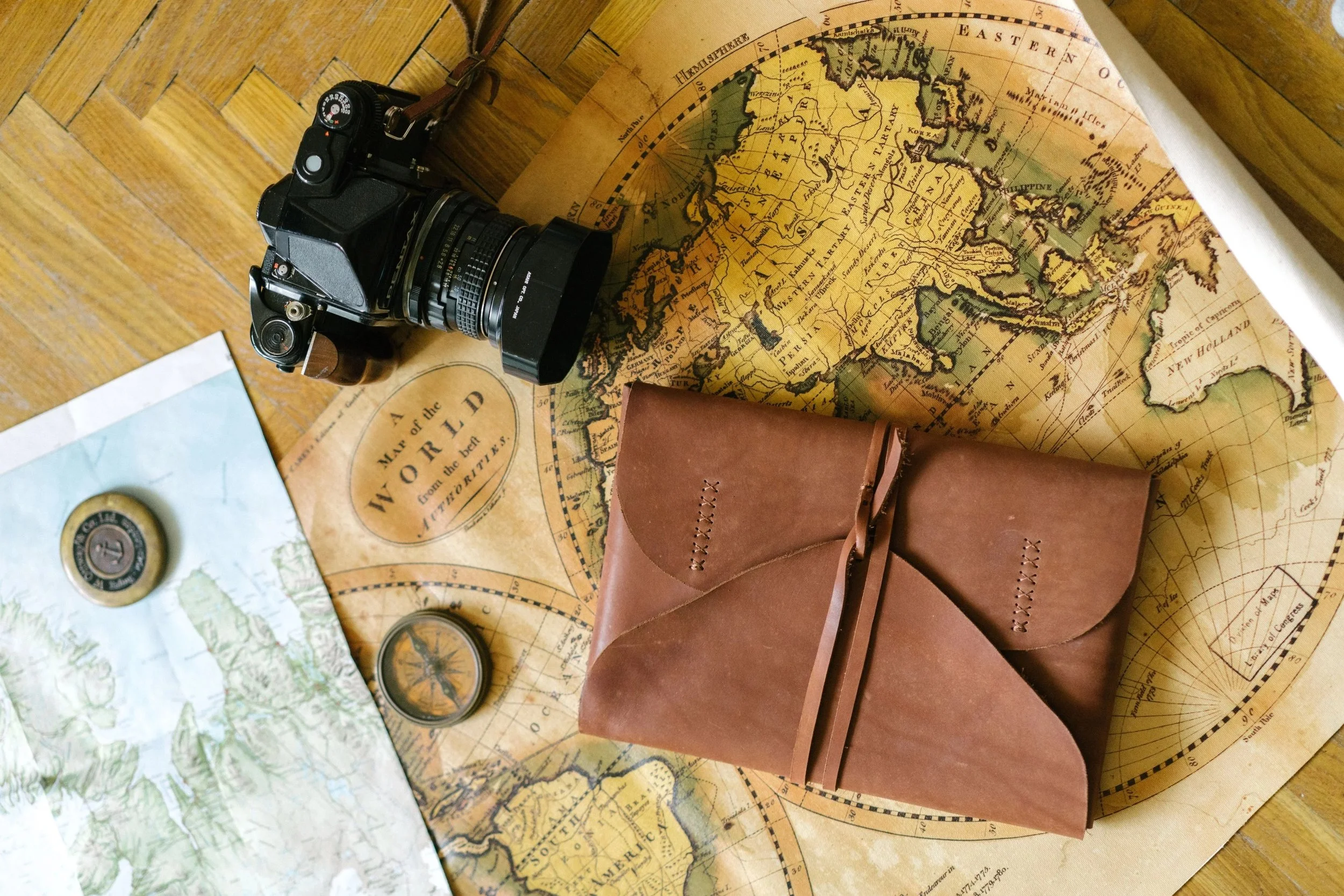 A vintage map of the world, a camera with a telephoto lens, two leather pouches, a compass, a pocket watch, and a smaller map are spread out on a wooden surface.