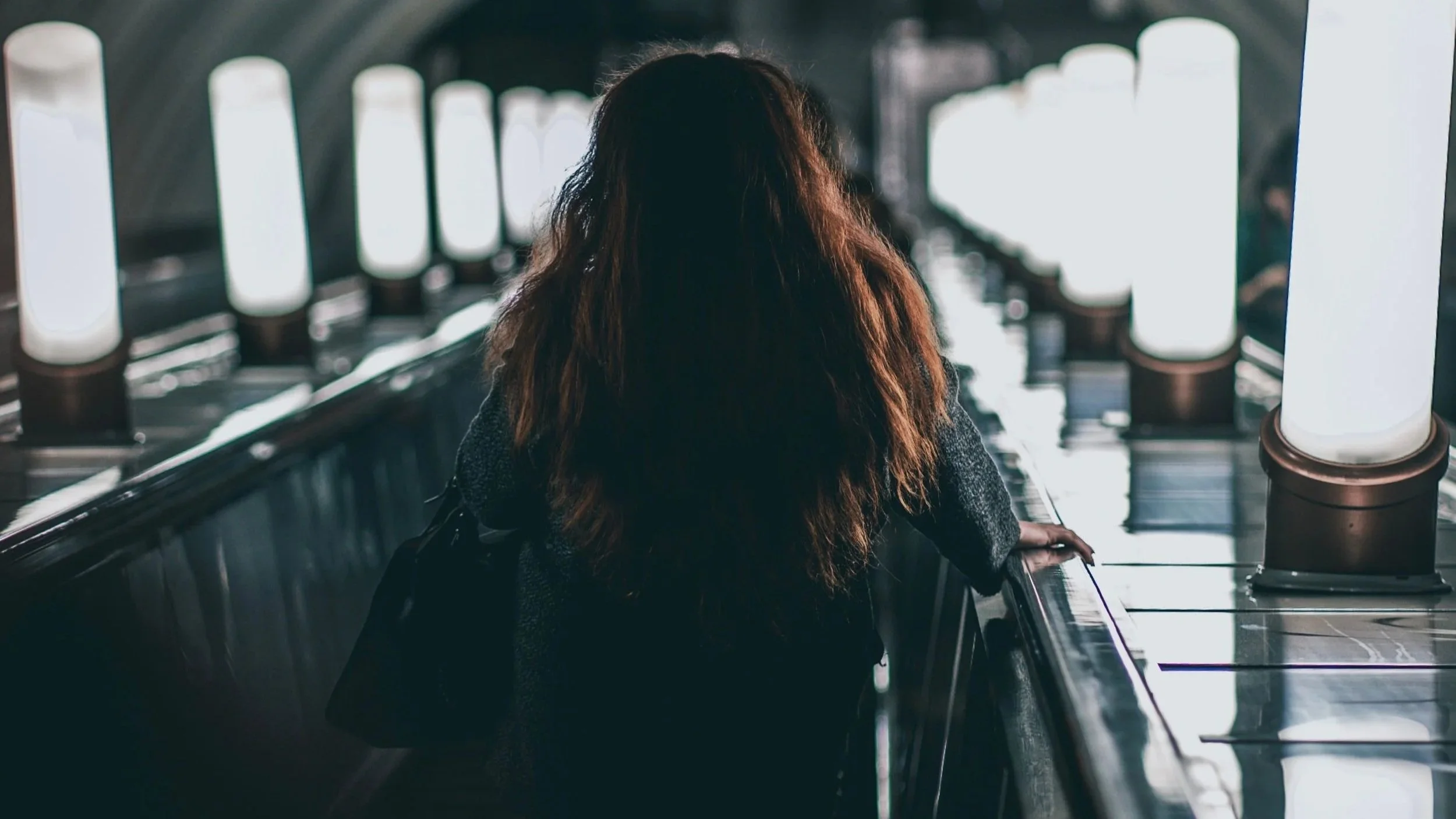 A person with long hair riding an escalator in an airport with bright lights.
