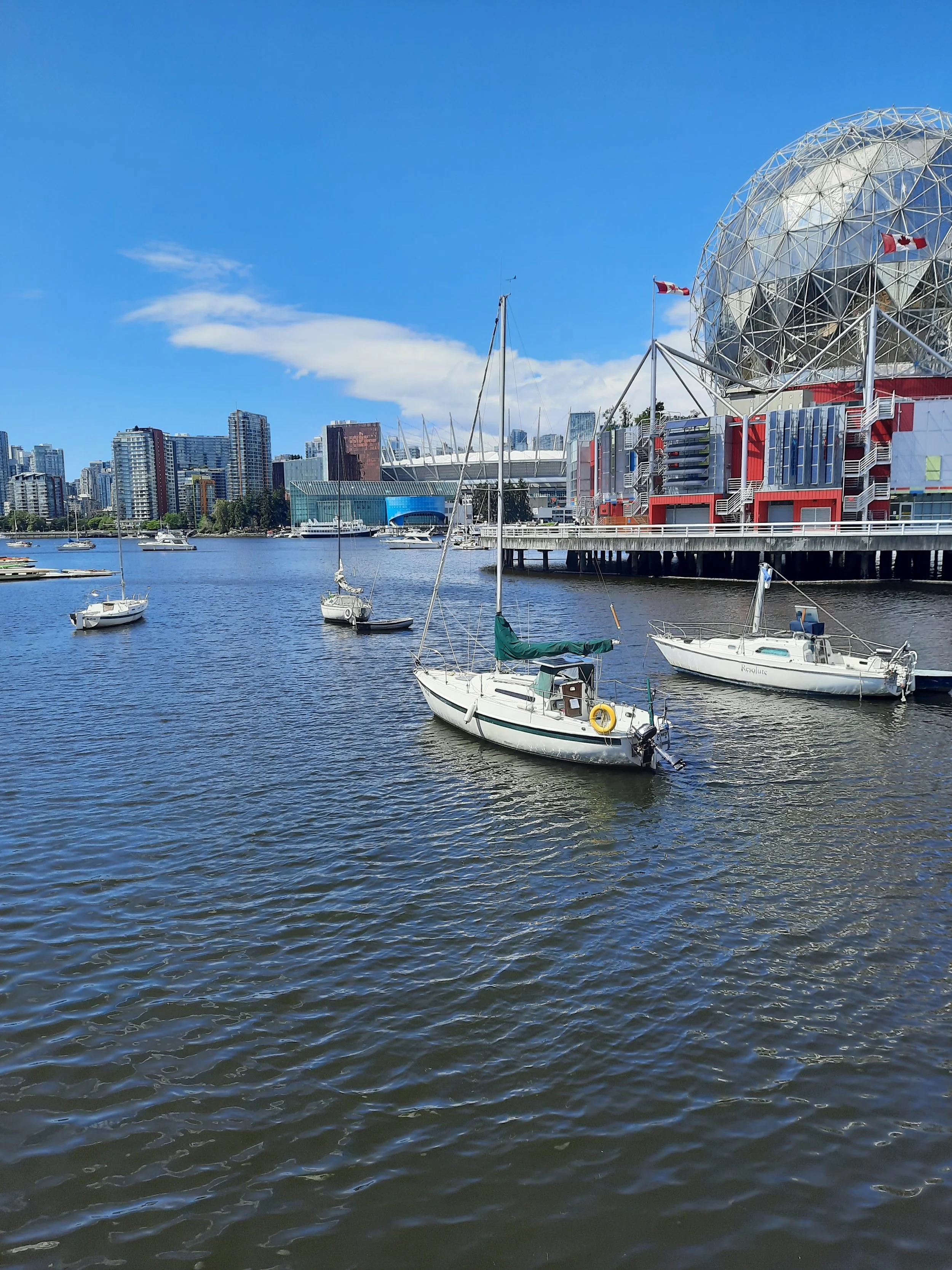 View of a harbor with sailboats on the water and modern high-rise buildings in the background, under a bright blue sky with some clouds in Vancouver British Columbia.