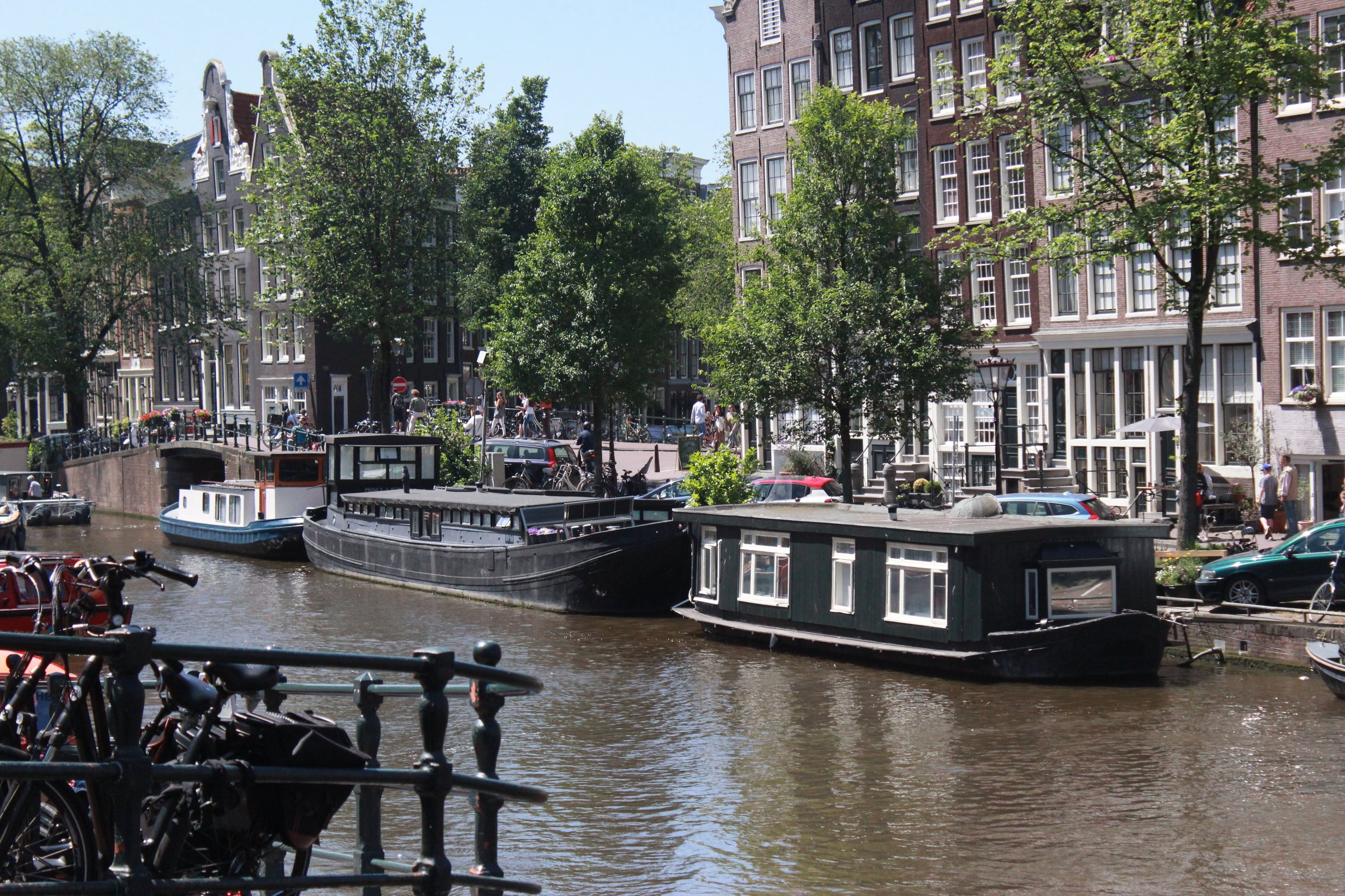 Canal with boats docked along the edge, historic brick buildings with large windows, trees, and a crowd of pedestrians walking on the street, sunny day in Amsterdam.