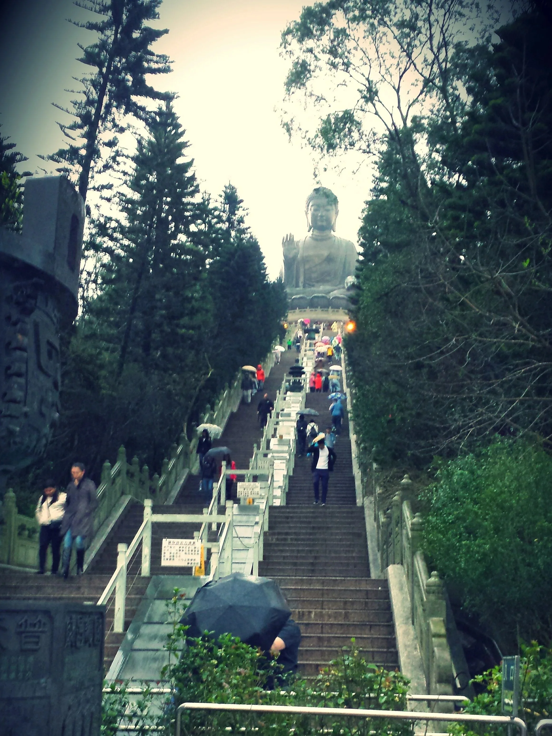 People walking up and down a long stairway flanked by trees, with a large Buddha statue at the top of the stairs and a bright sky in the background in Hong Kong.