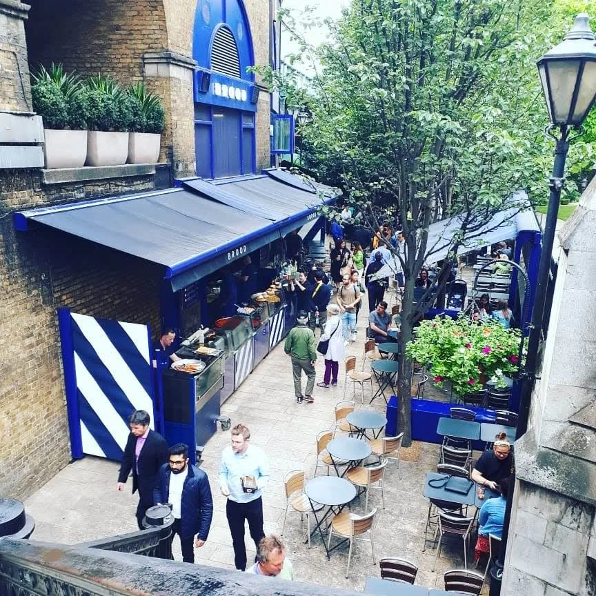 A bustling outdoor street café with tables and chairs, people walking and dining, a food stall with a blue awning, greenery with trees and potted plants, on a city street with brick buildings and a lamppost.