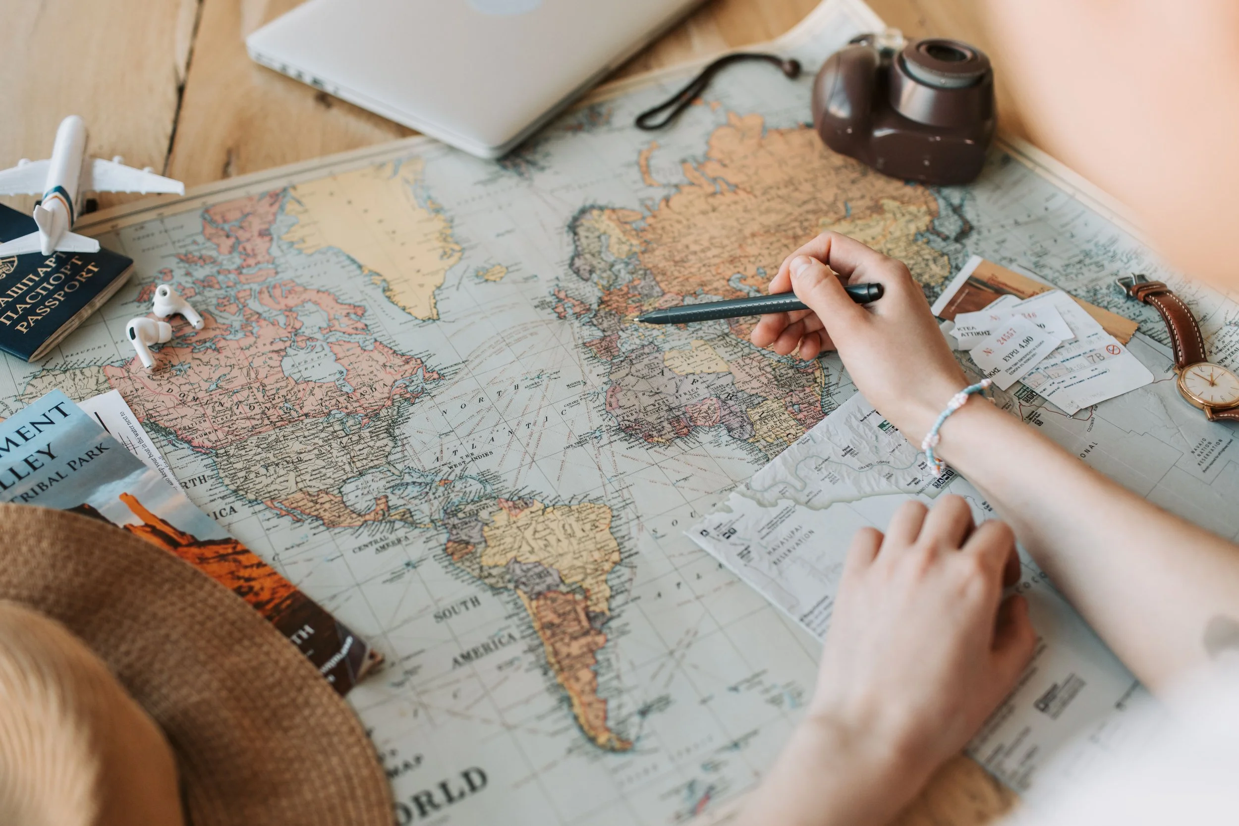Person marking a world map with a black pen, surrounded by travel documents, a camera, a watch, tickets, and an airplane model.