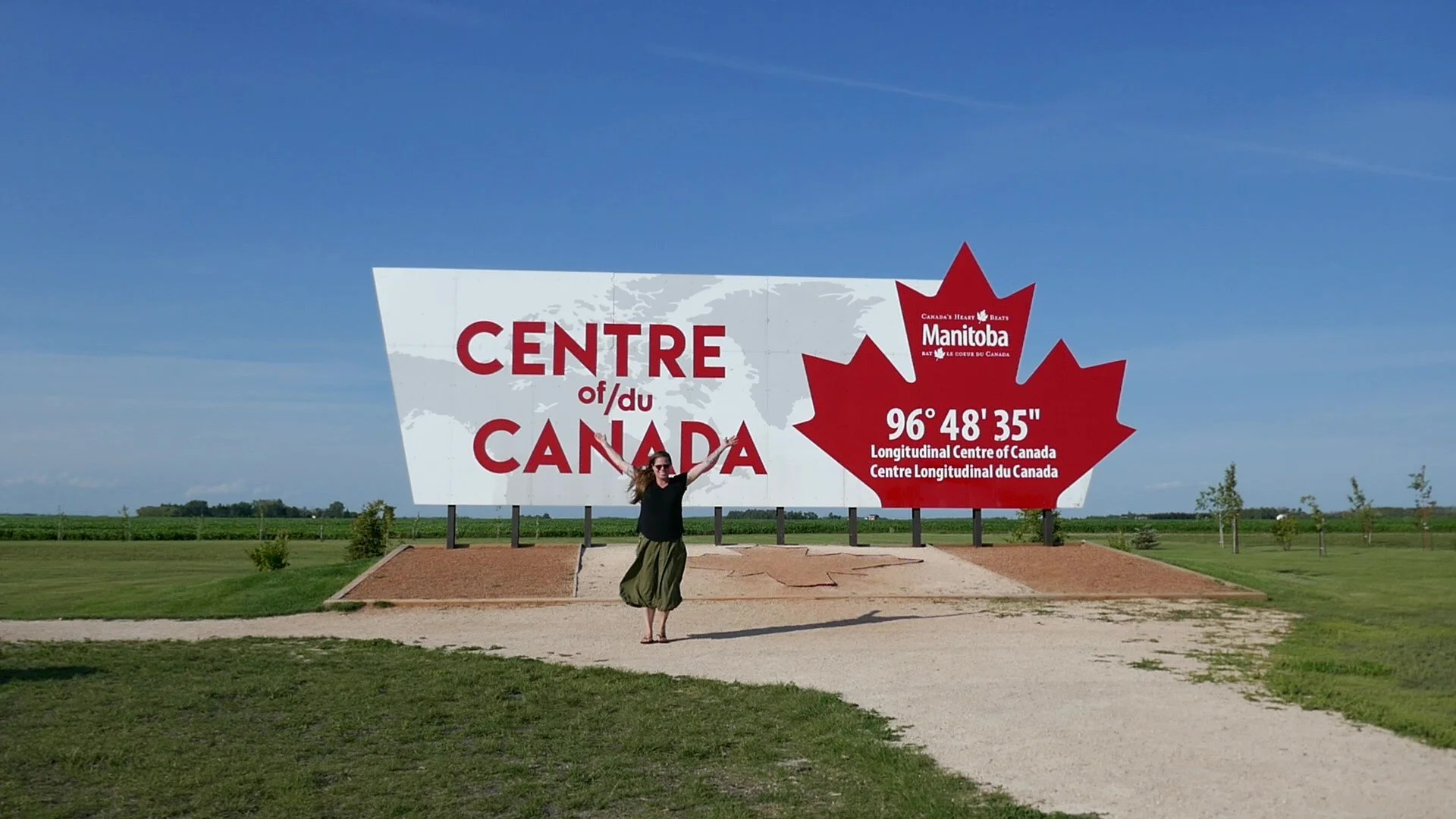 A woman standing in front of a large sign indicating the geographic center of Canada in Manitoba. The sign features a red maple leaf with the coordinates 96°48'35" and she is wearing sunglasses, a black top, and a green skirt, raising her arms.