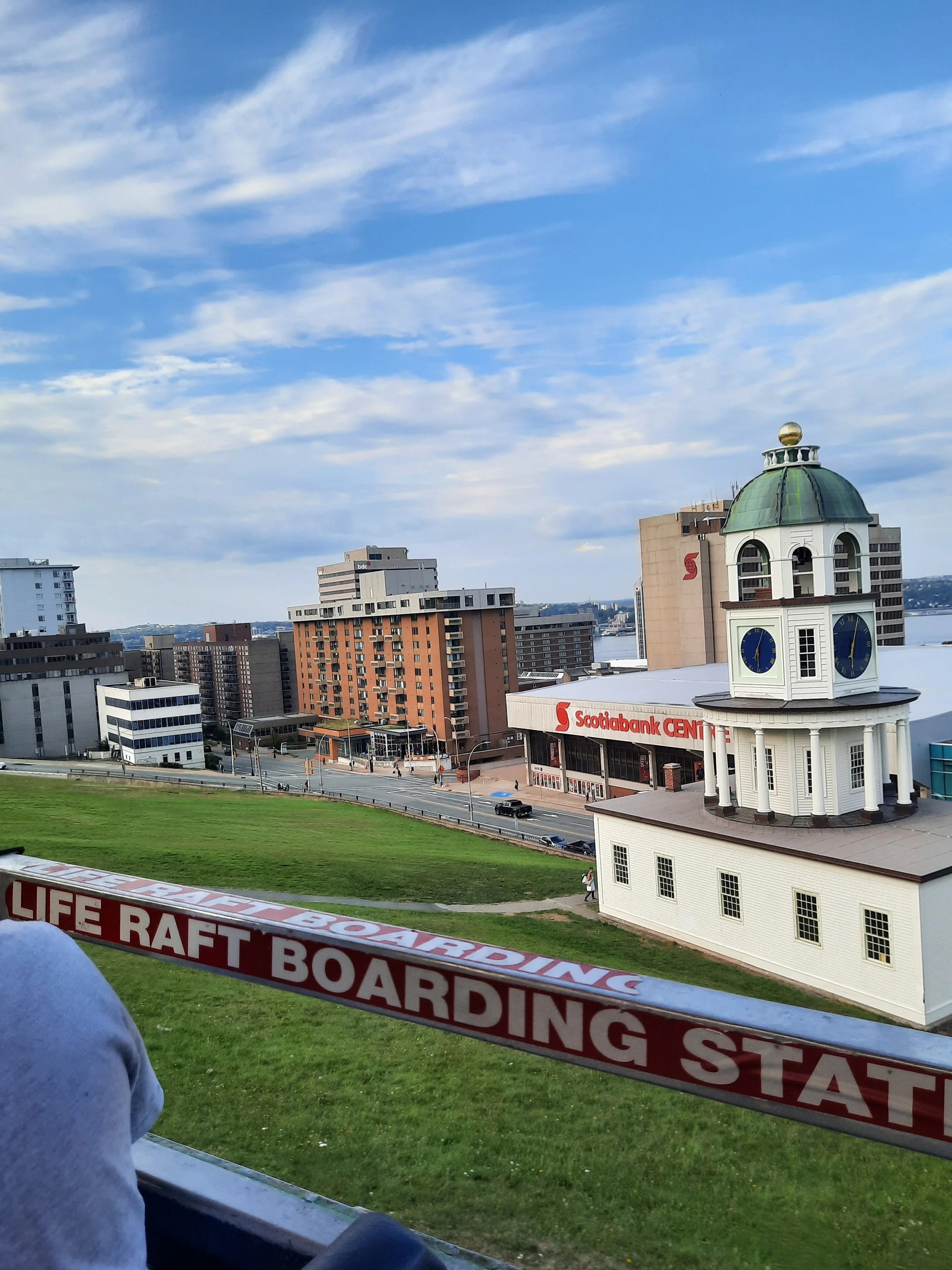Cityscape view showing a historic clock tower with a green dome, modern high-rise buildings, a river in the background, and a green park in the foreground. A sign in the foreground reads 'LIFE RAFT BOARDING'. The sky is partly cloudy in Halifax