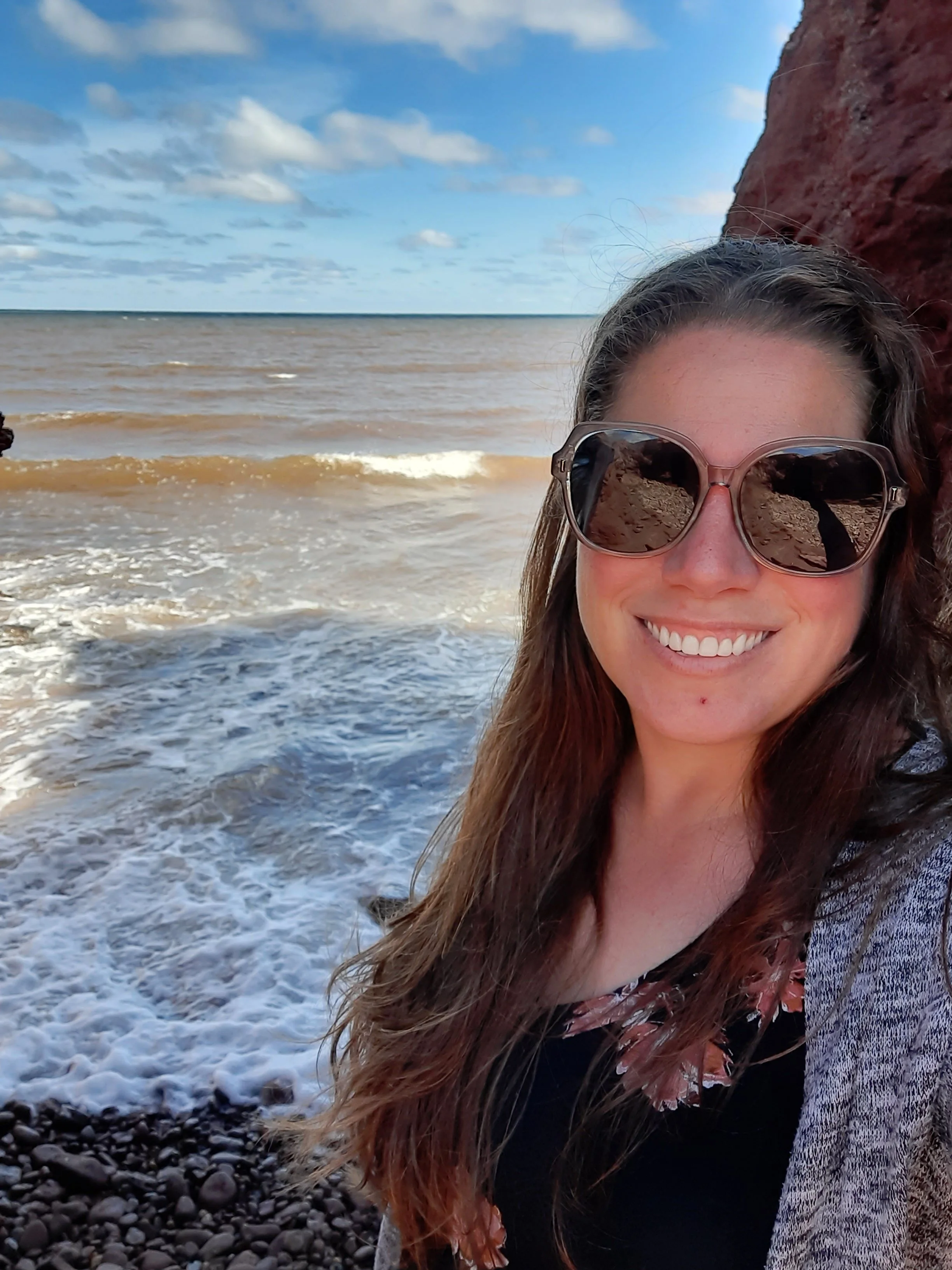 A woman smiling wearing sunglasses at a beach with waves and a cloudy sky in the background.