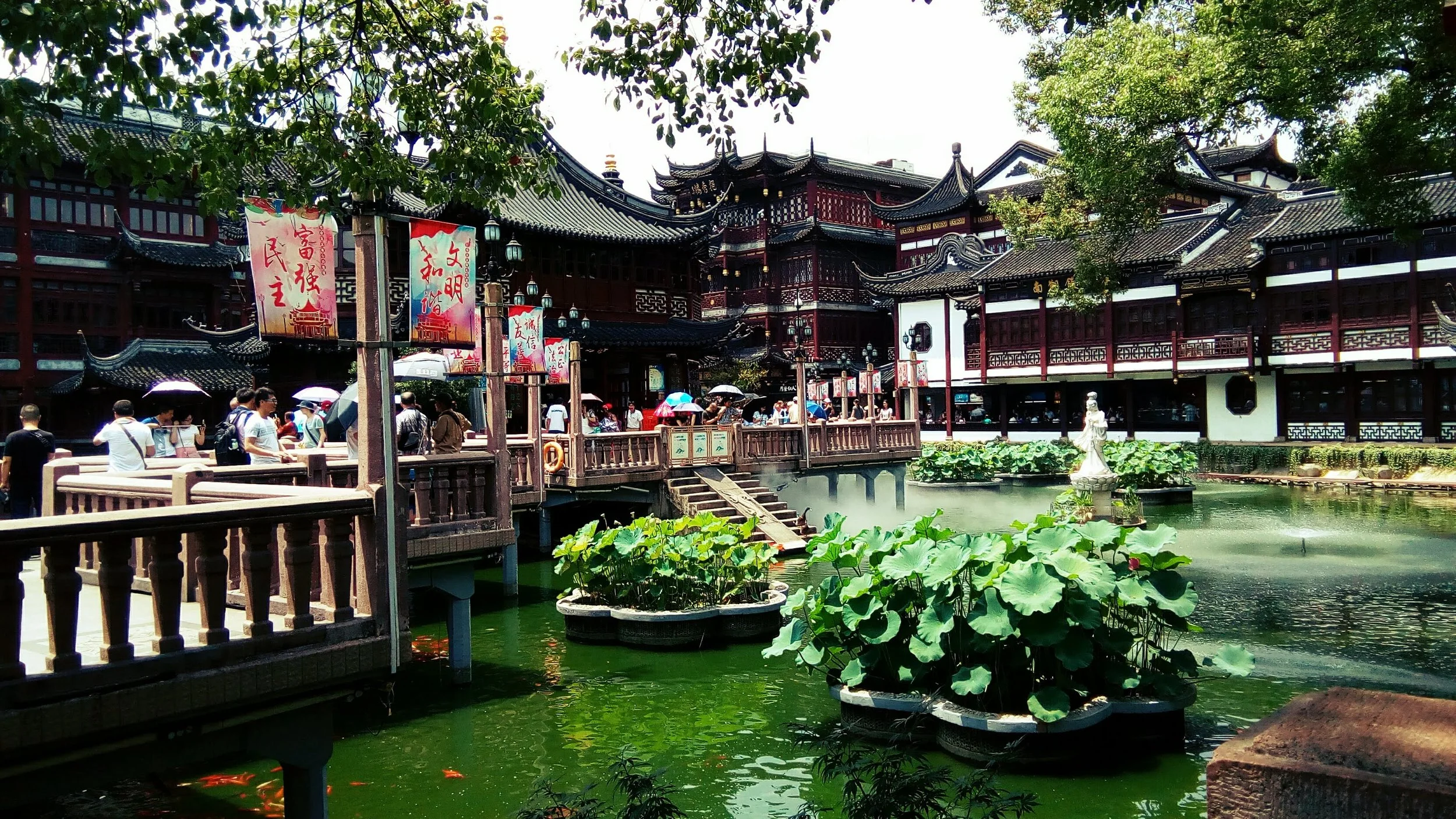 Traditional Chinese architectural buildings with dark tiled roofs and ornate wooden details surrounding a pond filled with lotus plants, with people walking on a wooden bridge and holding umbrellas in Yu Garden in Shanghai China.