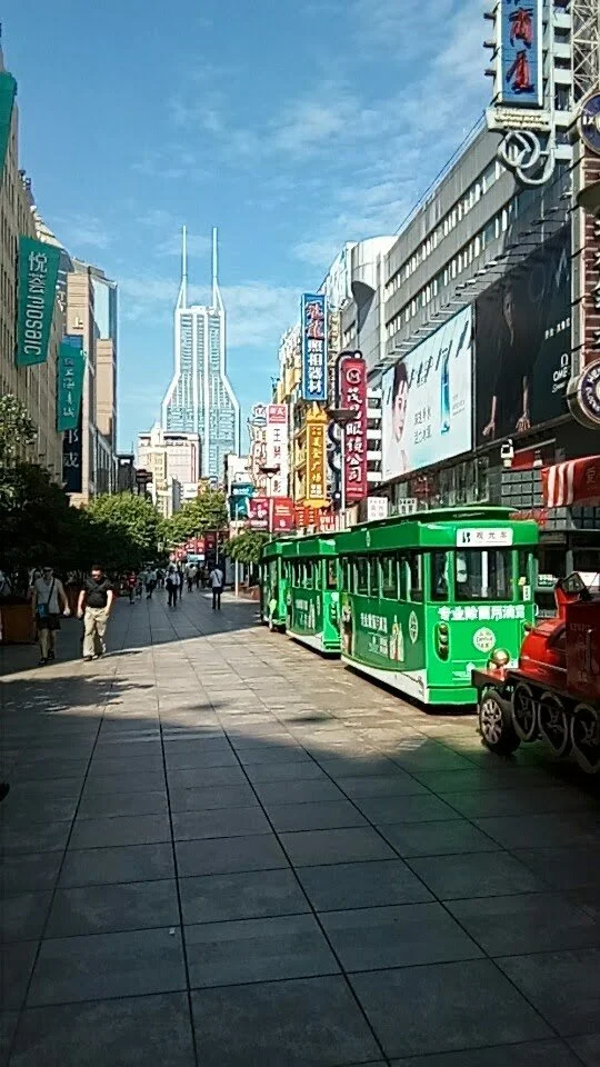 A busy city street with green trams, people walking, and tall buildings with colorful signs and billboards in the background in Shanghai China
