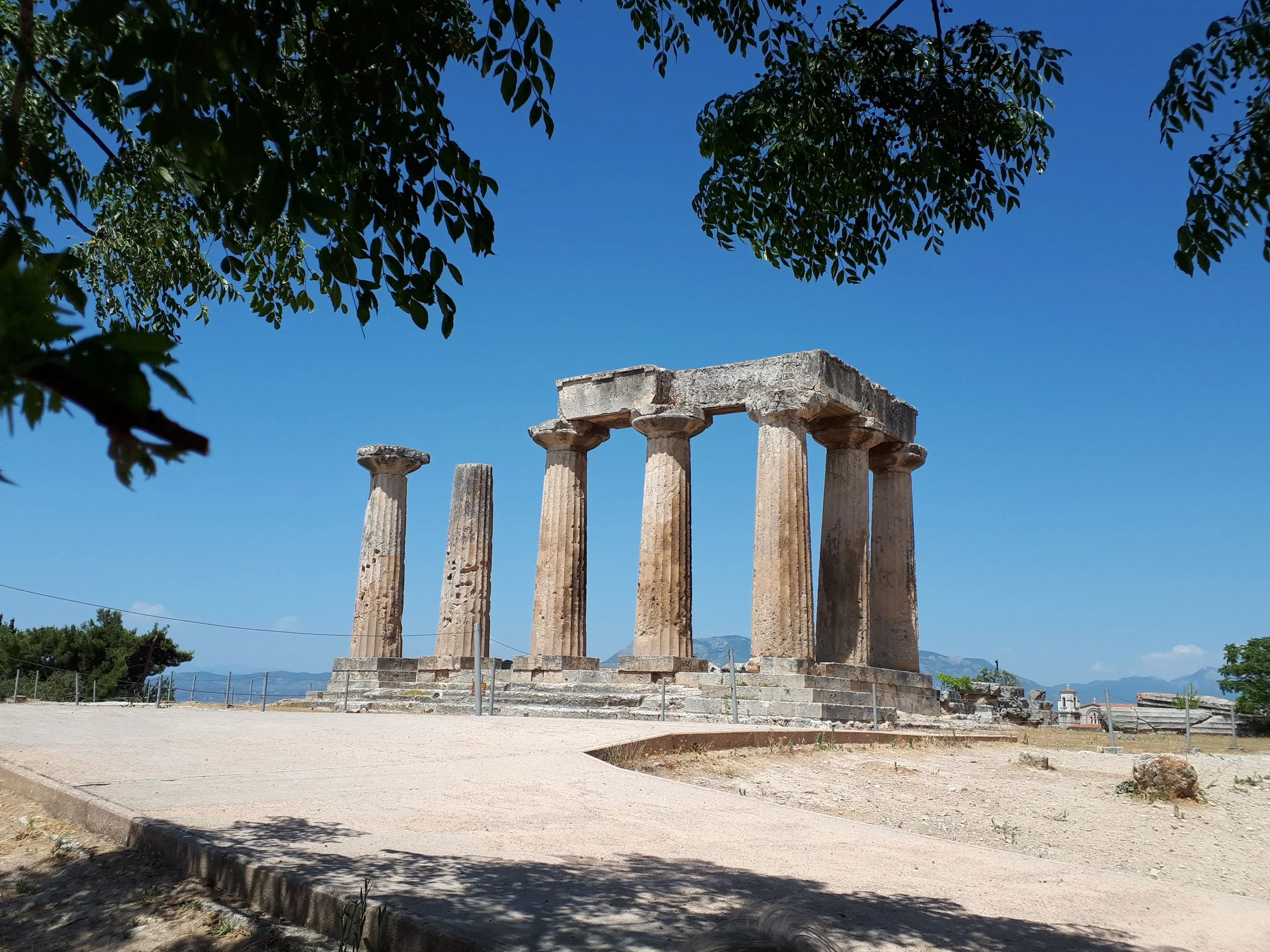 Ancient ruins with Greek-style columns, some topped with a stone structure, situated outdoors under a clear blue sky with green trees partially framing the image.