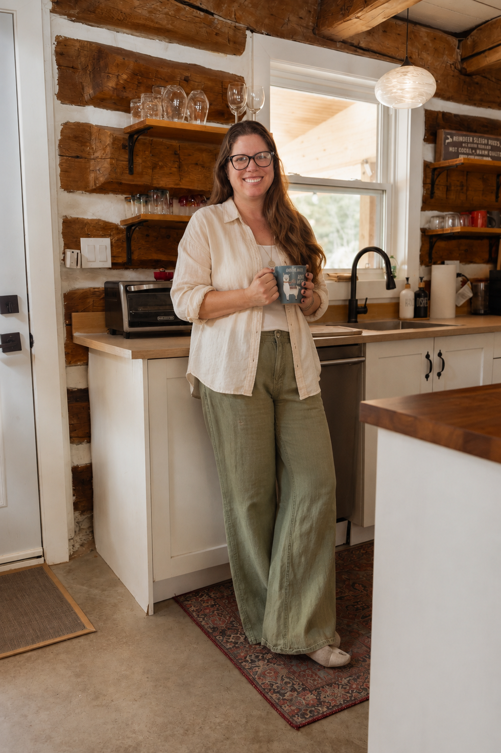 A woman with long brown hair and glasses smiling while holding a coffee mug in a cozy rustic kitchen with wooden beams and shelves.