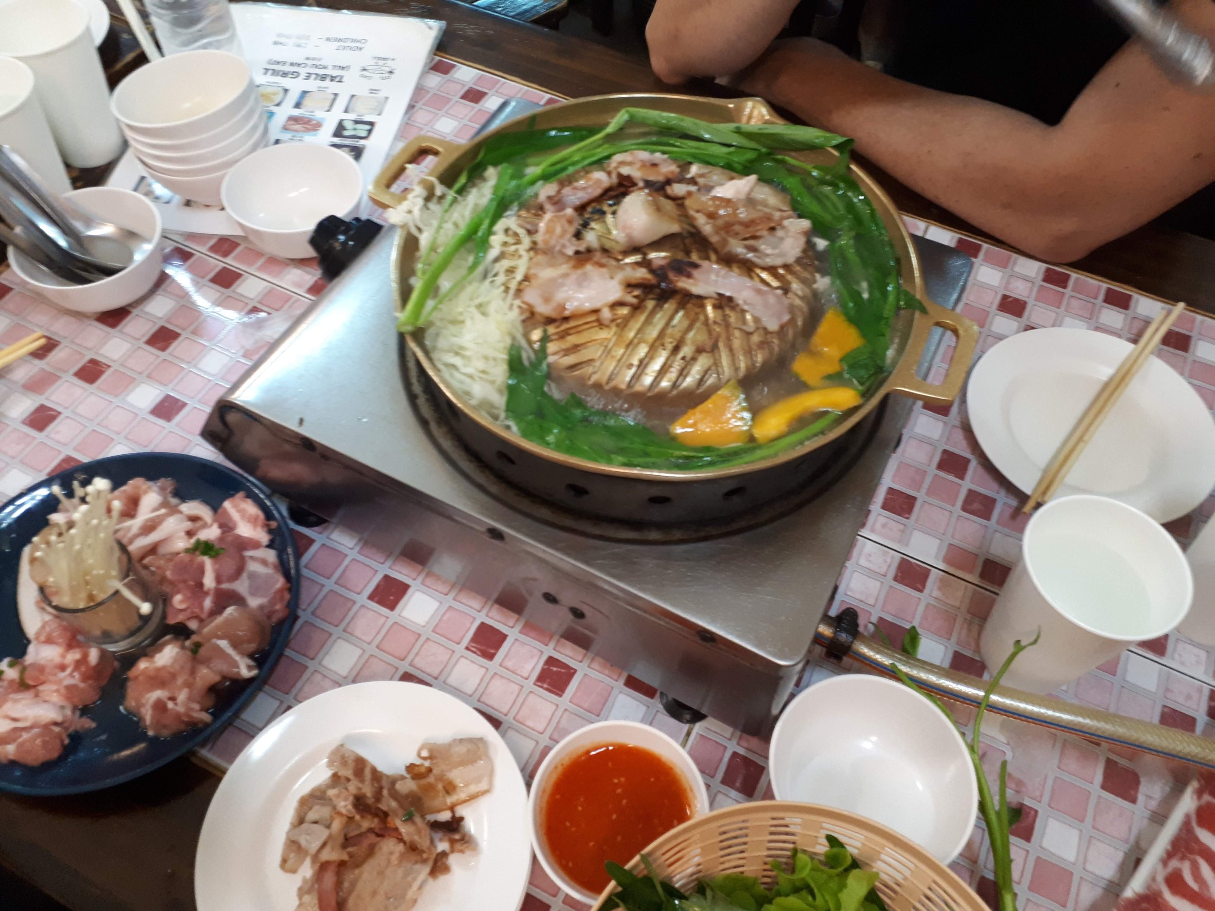 Mookata, Plate of grilled meat, green onions, and vegetables on a grill at a table with various small bowls of dipping sauces and additional plates of raw meat, chopsticks, and a condiment jar.