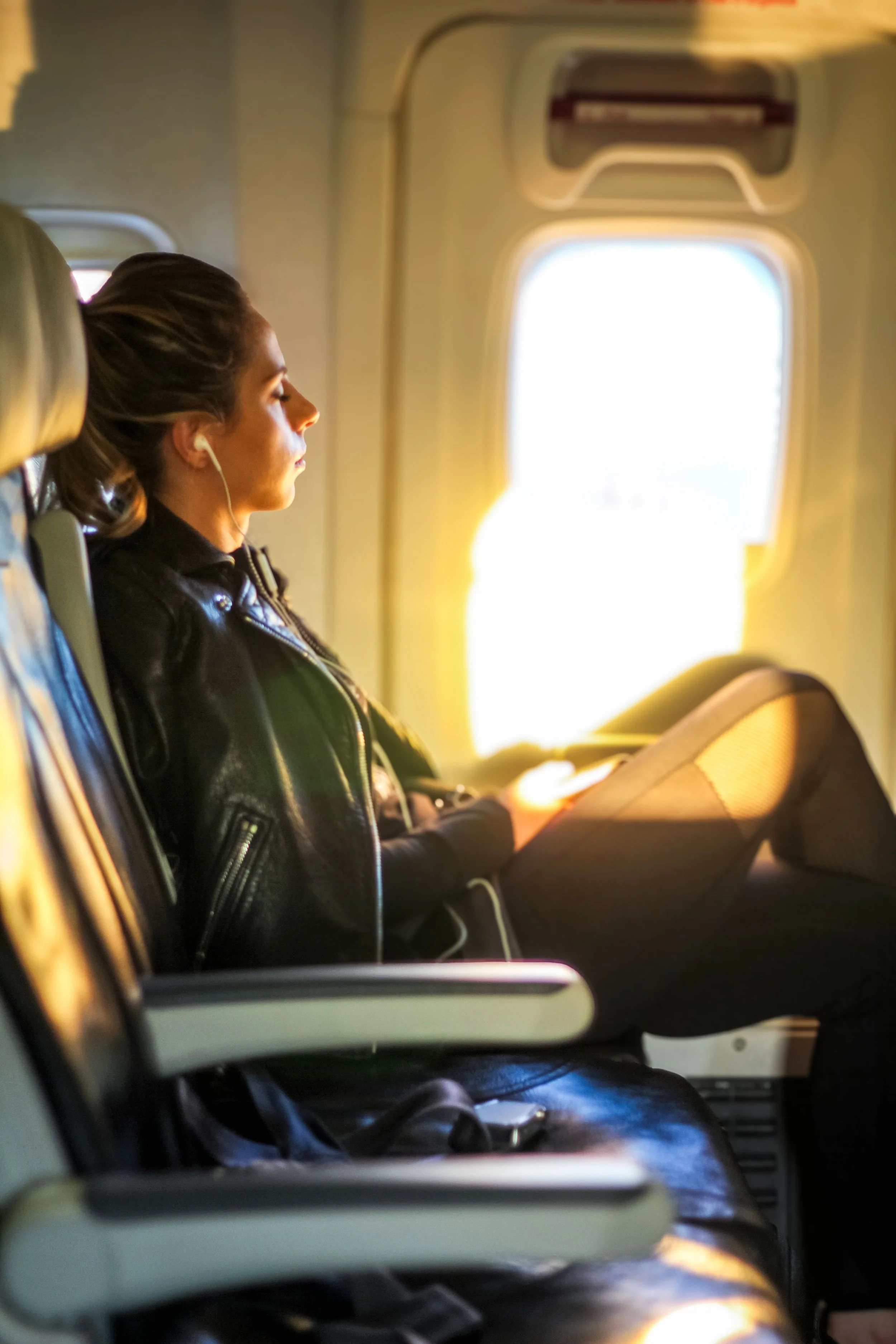 A woman sitting on an airplane, with her eyes closed, listening to music through earphones, and reading a book or magazine.