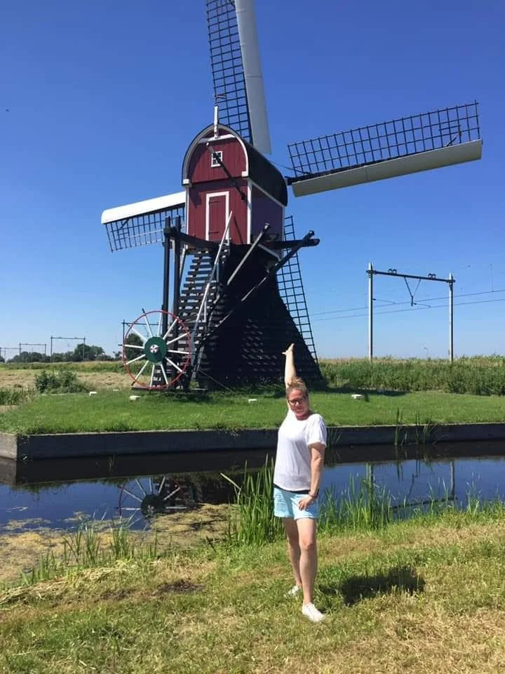 A woman standing next to a small pond with a windmill sculpture in the background. The windmill is red with black blades, and the sky is clear and blue.