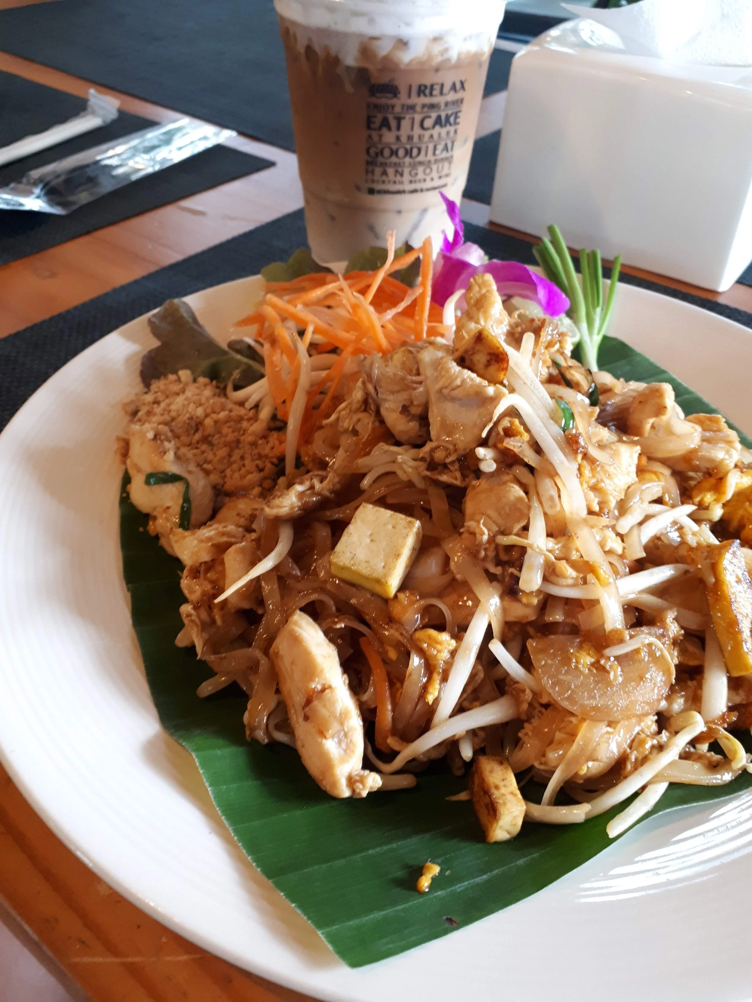 A plate of Pad Thai consisting of stir-fried noodles with vegetables and tofu, garnished with purple flowers, served on a banana leaf, with a plastic cup of iced coffee in the background.