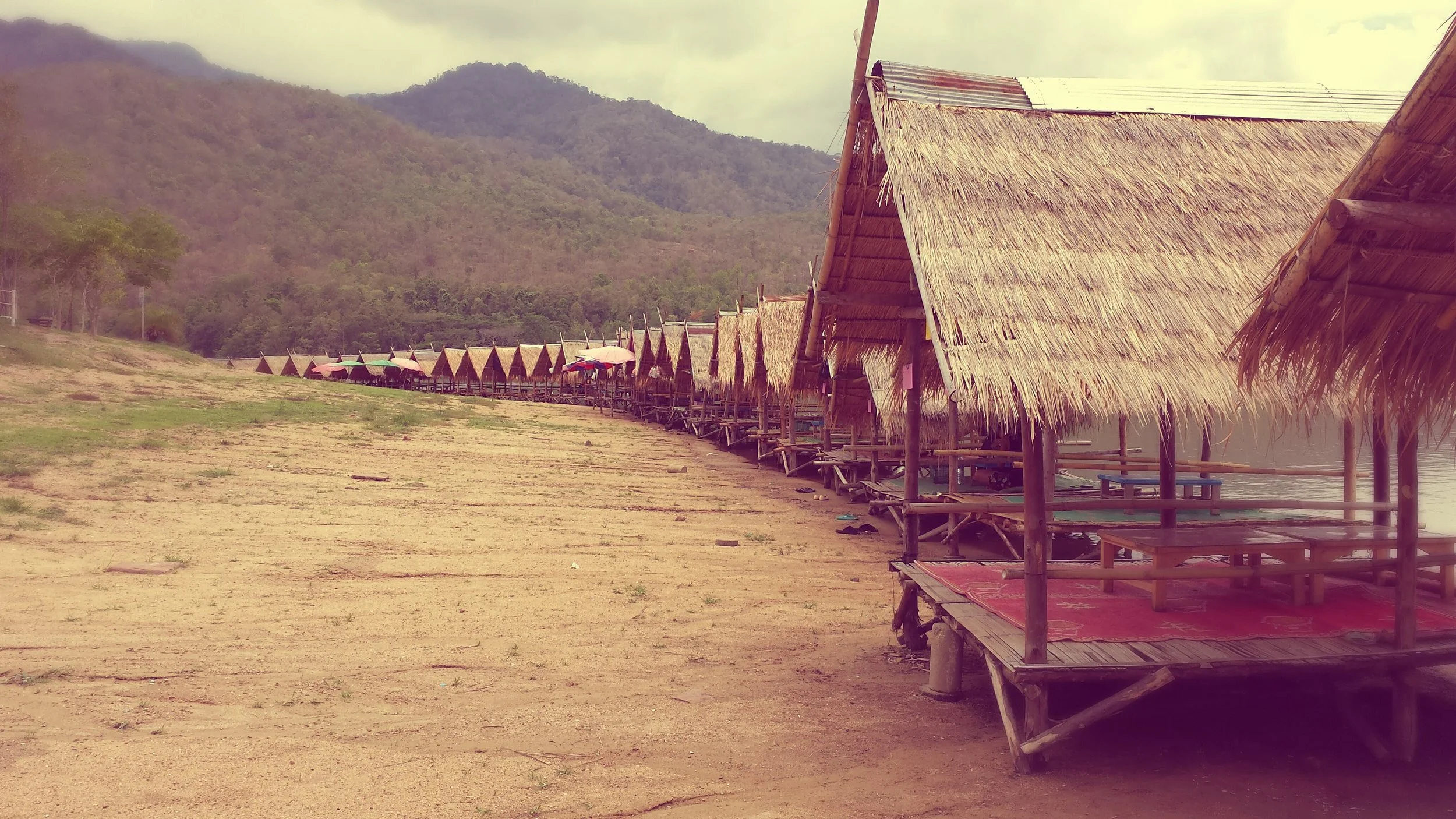 A row of thatched-roof huts built on stilts along a shoreline, with mountains in the background and umbrellas shading some huts in Chiang Mai Thailand.