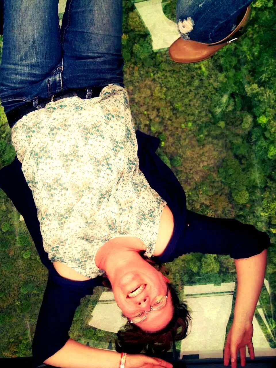 A woman with glasses and a patterned top lying upside down, seemingly on a glass floor, with outdoor greenery below, and a building structure visible in the background.