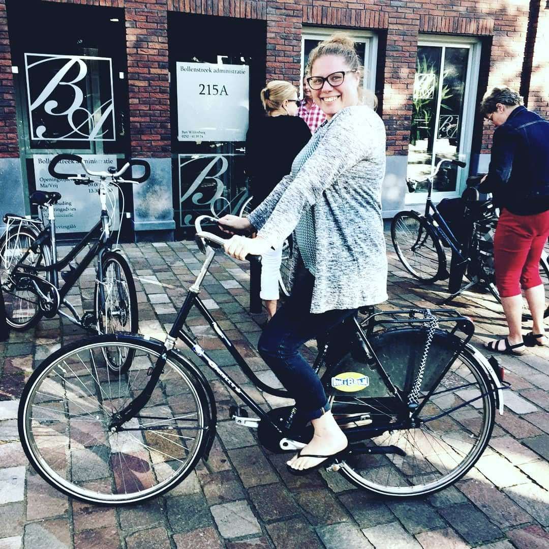 A smiling woman with glasses and a gray cardigan sitting on a black bicycle on a cobblestone street. Several people and bicycles are in the background outside a building with brick walls and glass windows.