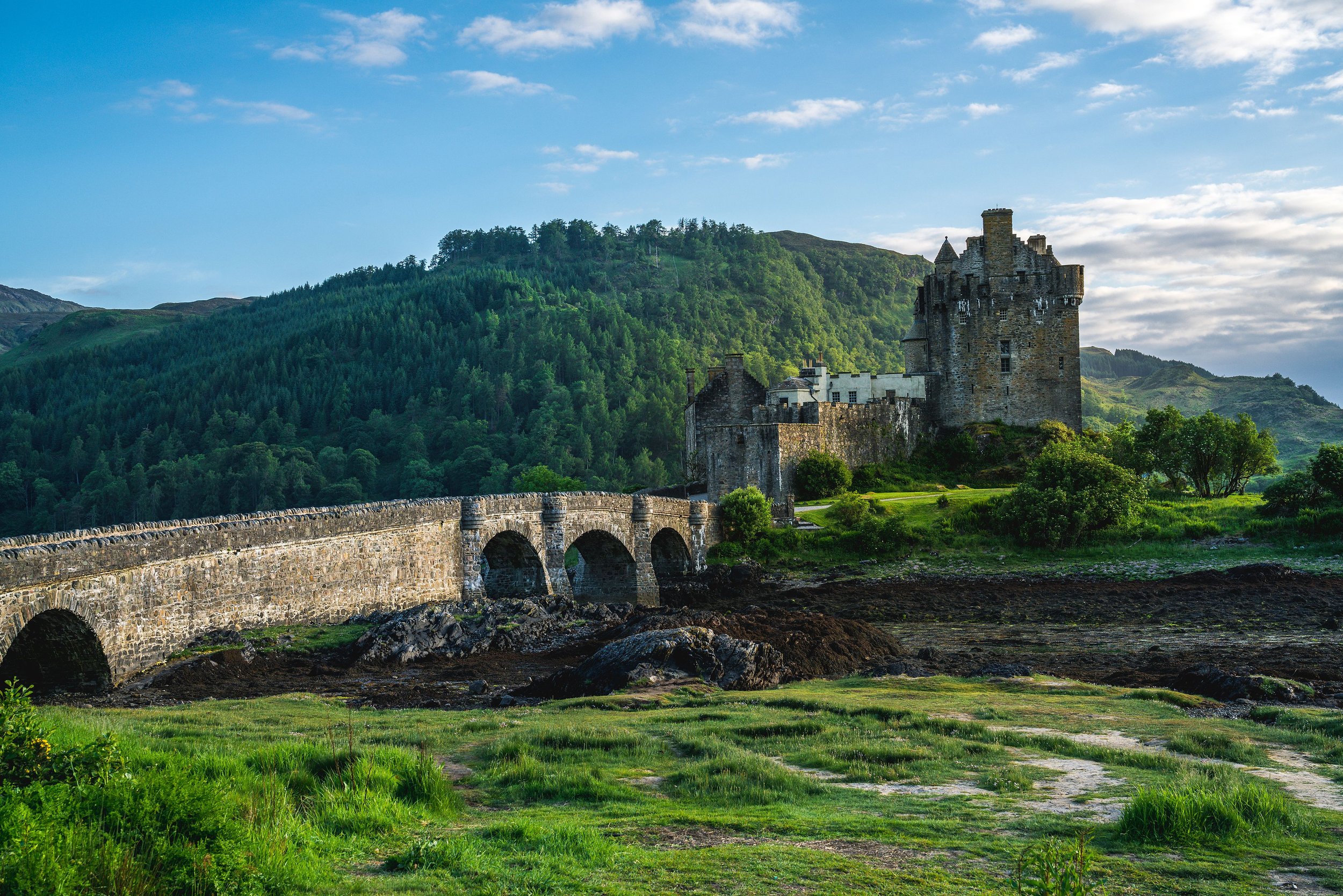 A historic castle on a hill with a stone bridge in the foreground, surrounded by lush green trees and hills under a partly cloudy sky.