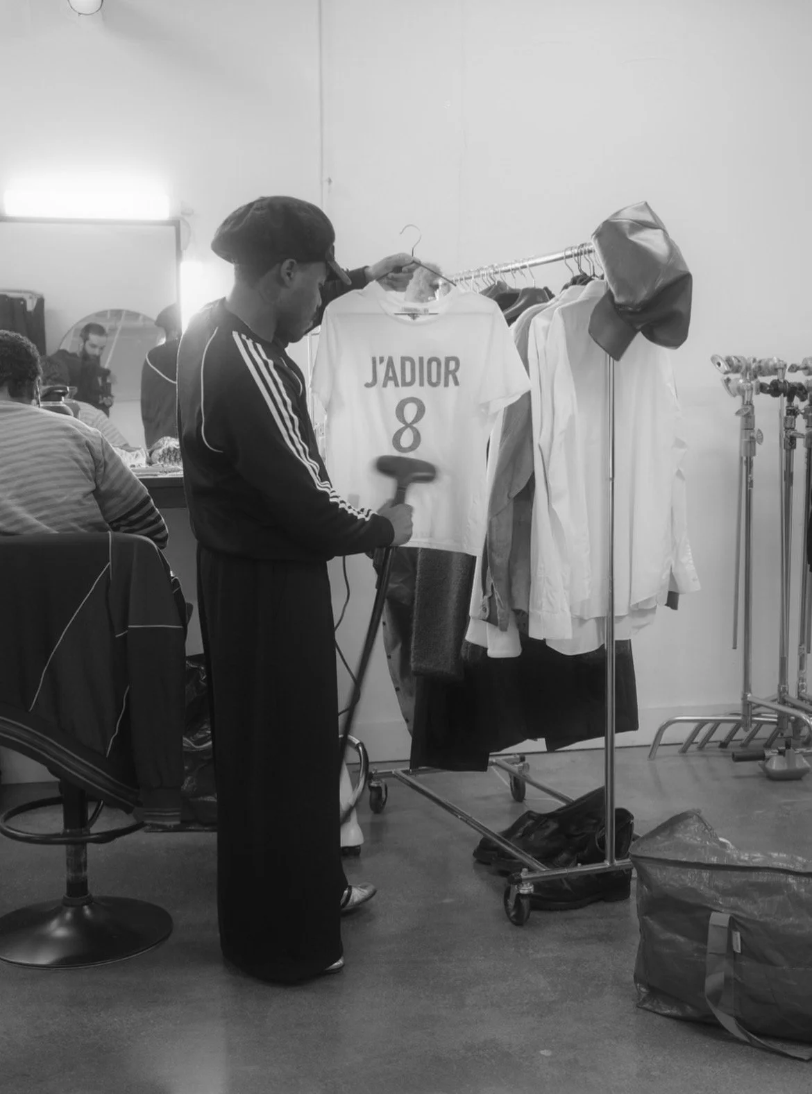 A man ironing a white Fendi T-shirt with J'Adior 8 printed on it, hanging on a clothing rack in a studio or backstage setting, surrounded by other clothing and bags.