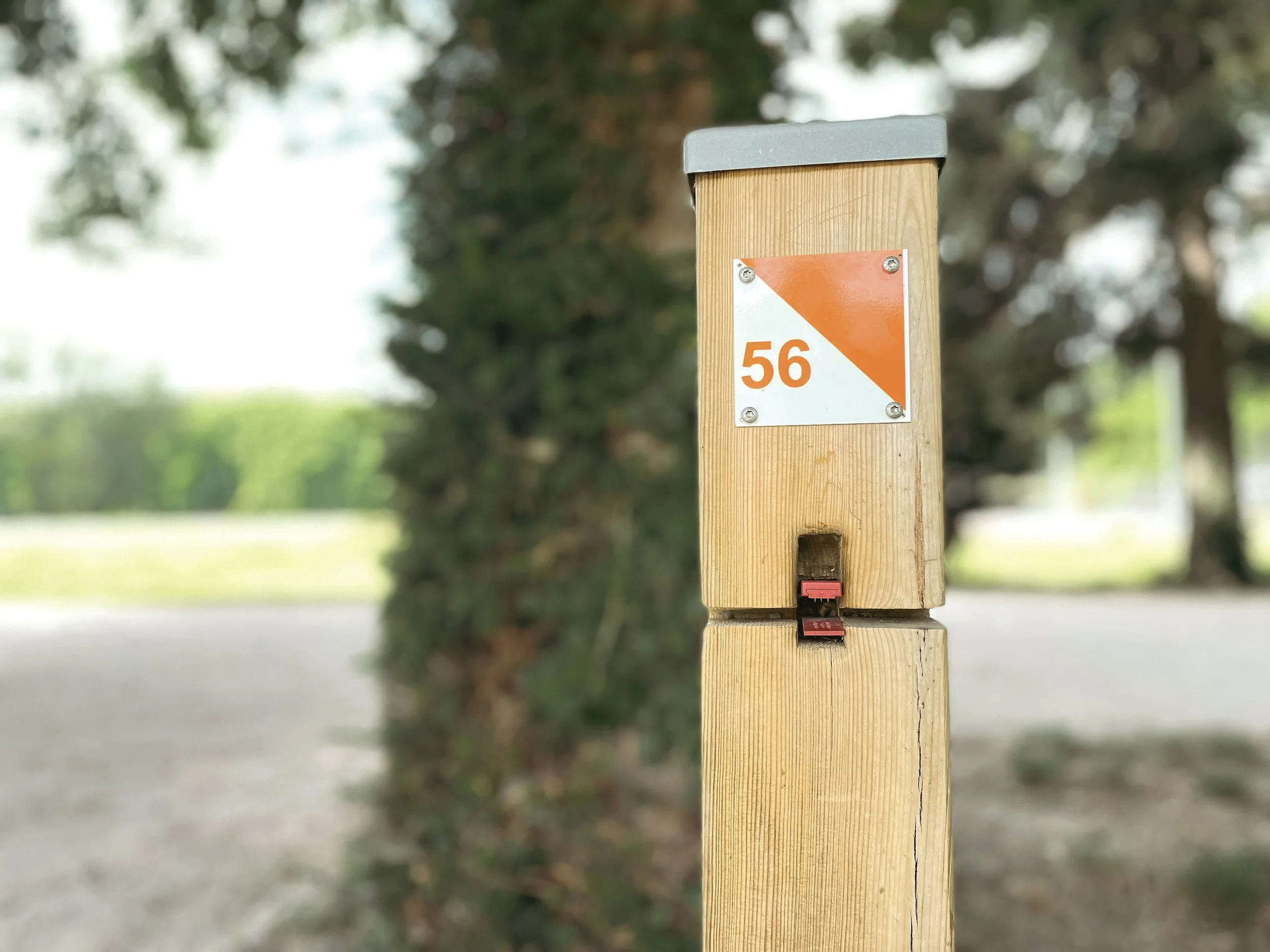 Un poteau en bois avec un panneau orange et blanc portant le numéro 56, située en plein air avec des arbres en arrière-plan.