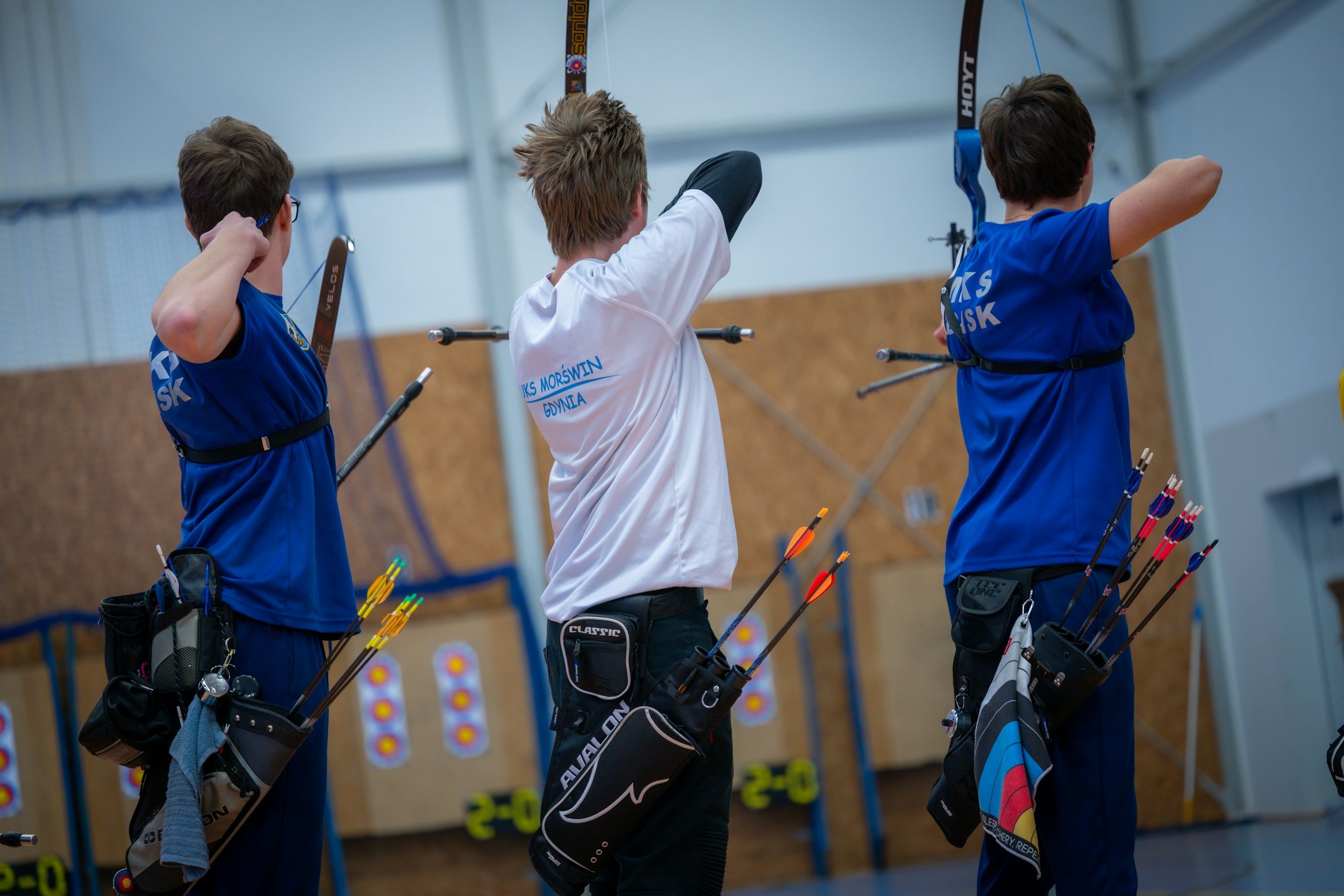 Trois jeunes archers sont alignés et en position de tir dans une salle d'arc. Ils portent des vêtements sportifs bleus et une chemise blanche, avec des équipements d'archerie, y compris des carquois remplis de flèches, fixés à leur taille.
