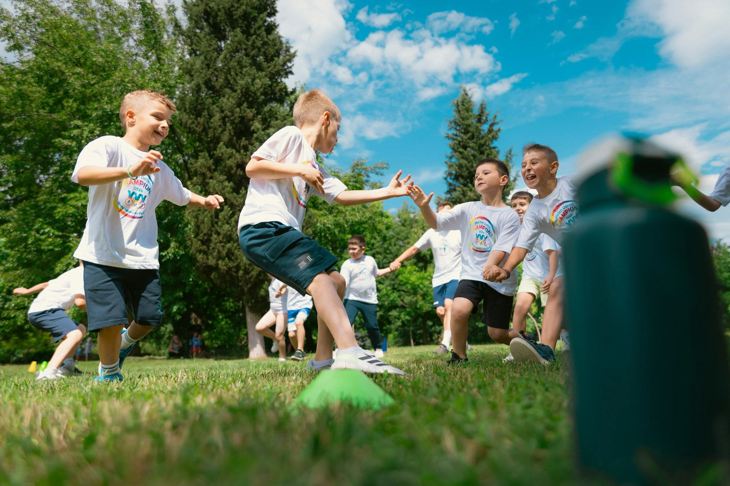 Des enfants jouent et s'amusent lors d'une activité en plein air dans un parc, sous un ciel bleu avec des nuages, portant tous des t-shirts blancs avec un logo coloré.