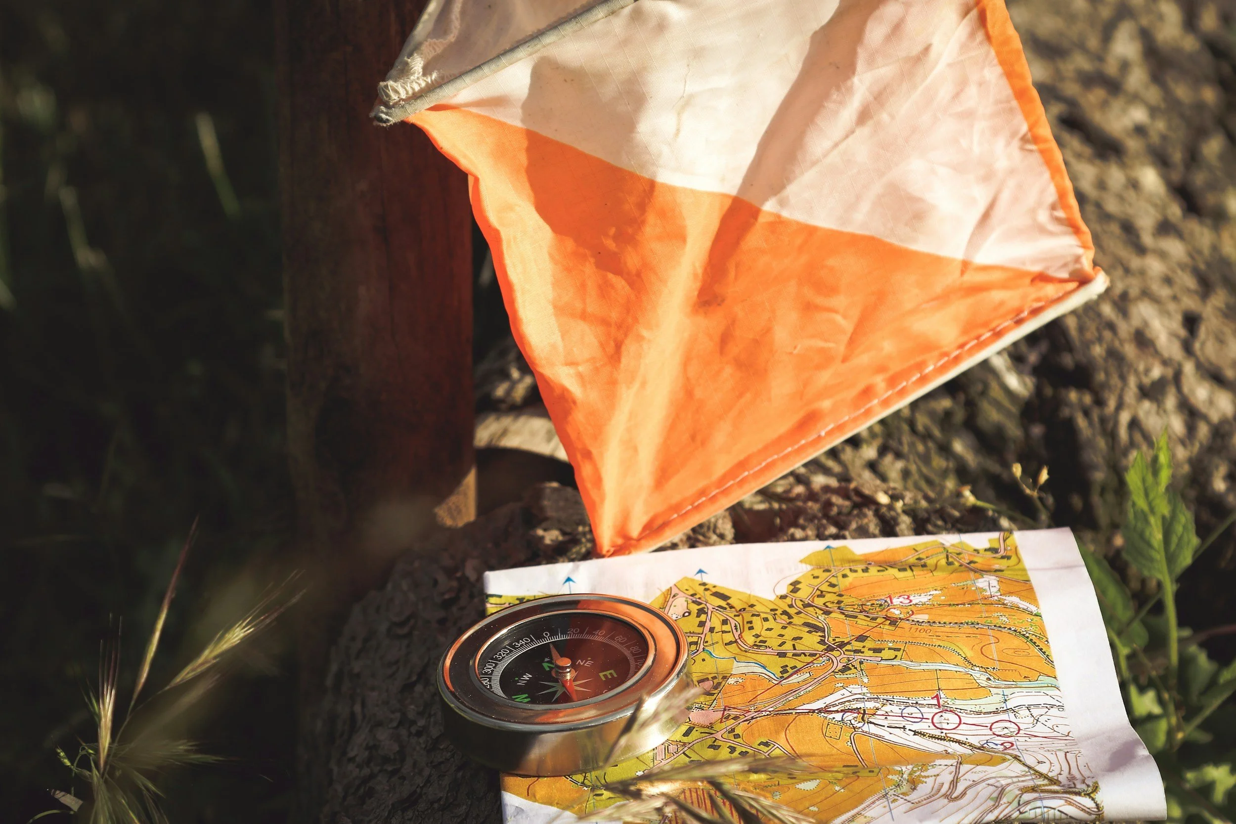 Une boussole, une carte et un drapeau orange et blanc sont posés sur le sol en terre, évoquant une activité de randonnée ou de navigation en plein air.