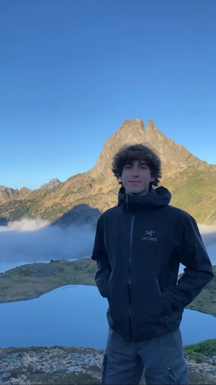 Jeune homme portant une veste noire, debout devant un lac de montagne avec une grande montagne derrière, ciel clair et beaux paysages alpins.