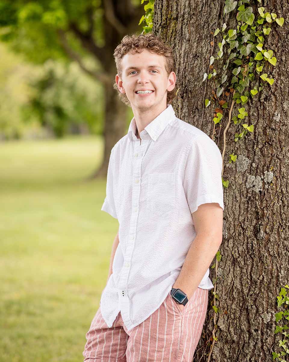 Smiling young man in a white shirt and striped shorts leaning against an ivy-covered tree outdoors