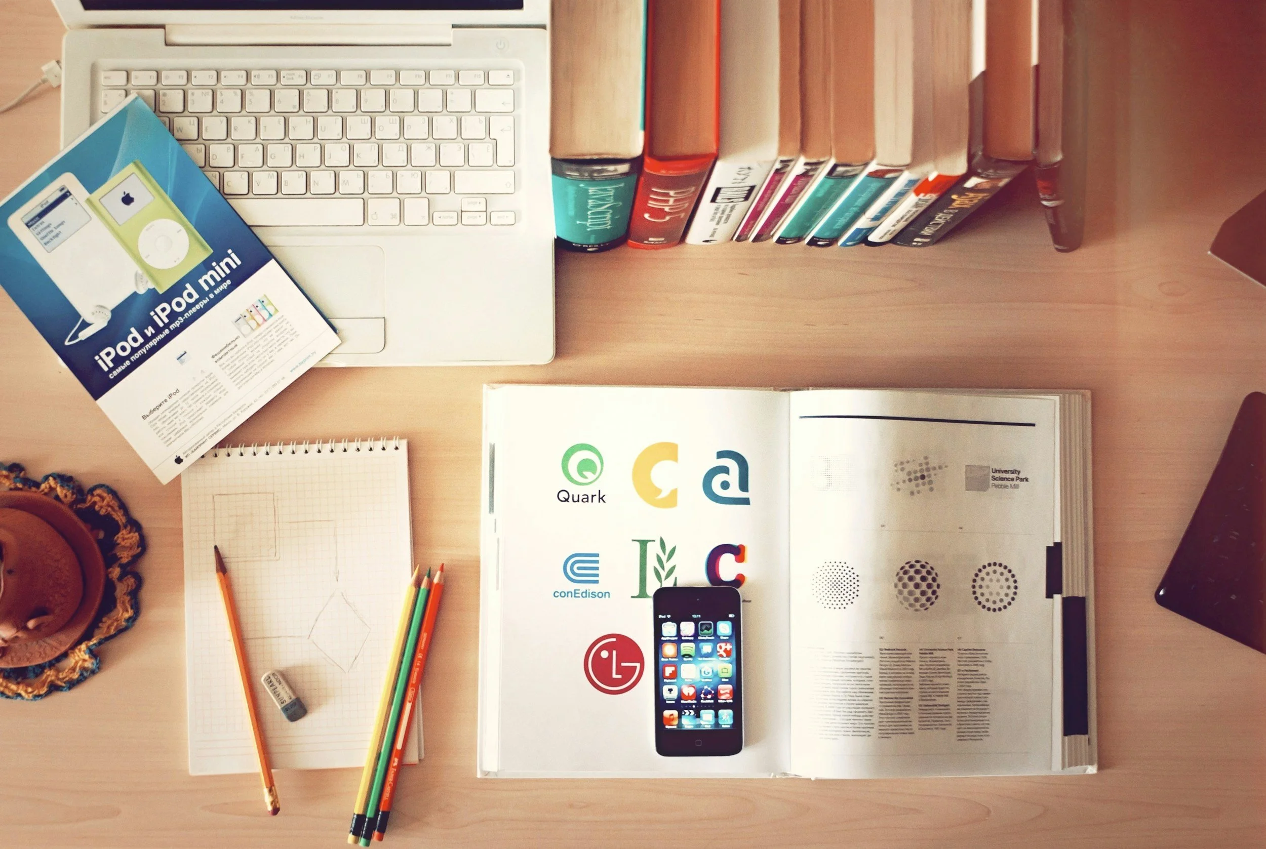 A desk with an open laptop, books, a pamphlet about iPod and iPhone, colored pencils, a spiral notepad, a magazine about science, a smartphone, and a cake.