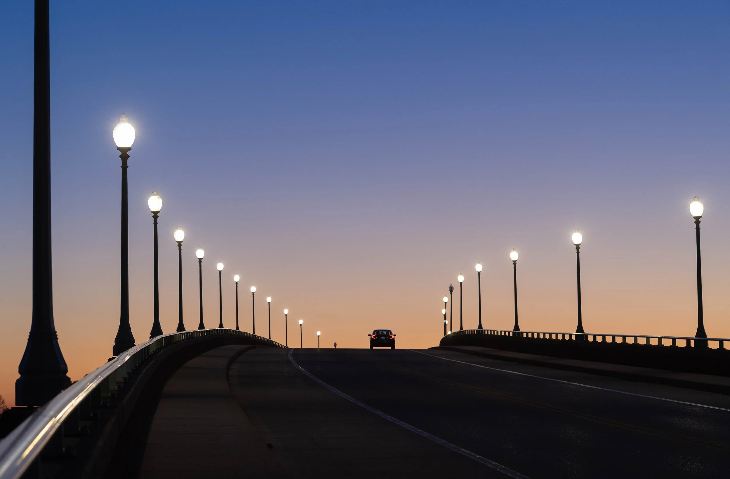 Bridge lined with illuminated vintage streetlamps at sunset, with a car driving toward the crest.