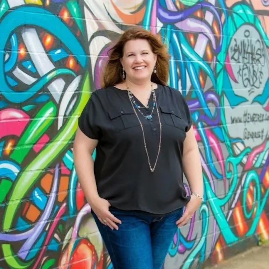 A smiling woman standing in front of a colorful graffiti mural, wearing a black blouse and blue jeans.