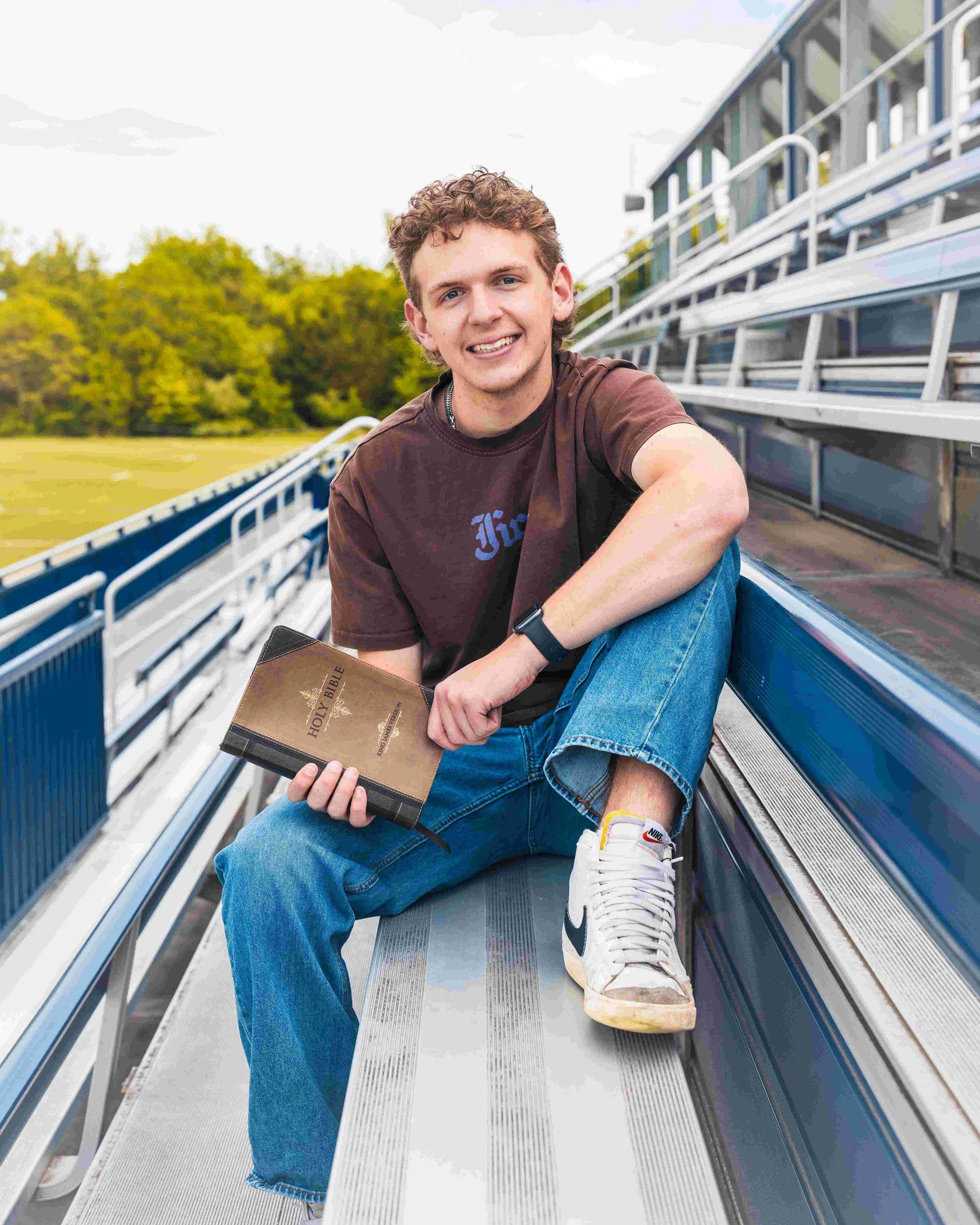 A young man with curly hair sitting on metal bleachers outdoors, holding a Bible with a camouflage cover, wearing a brown T-shirt, blue jeans, and white Nike sneakers, smiling at the camera, with green trees in the background.