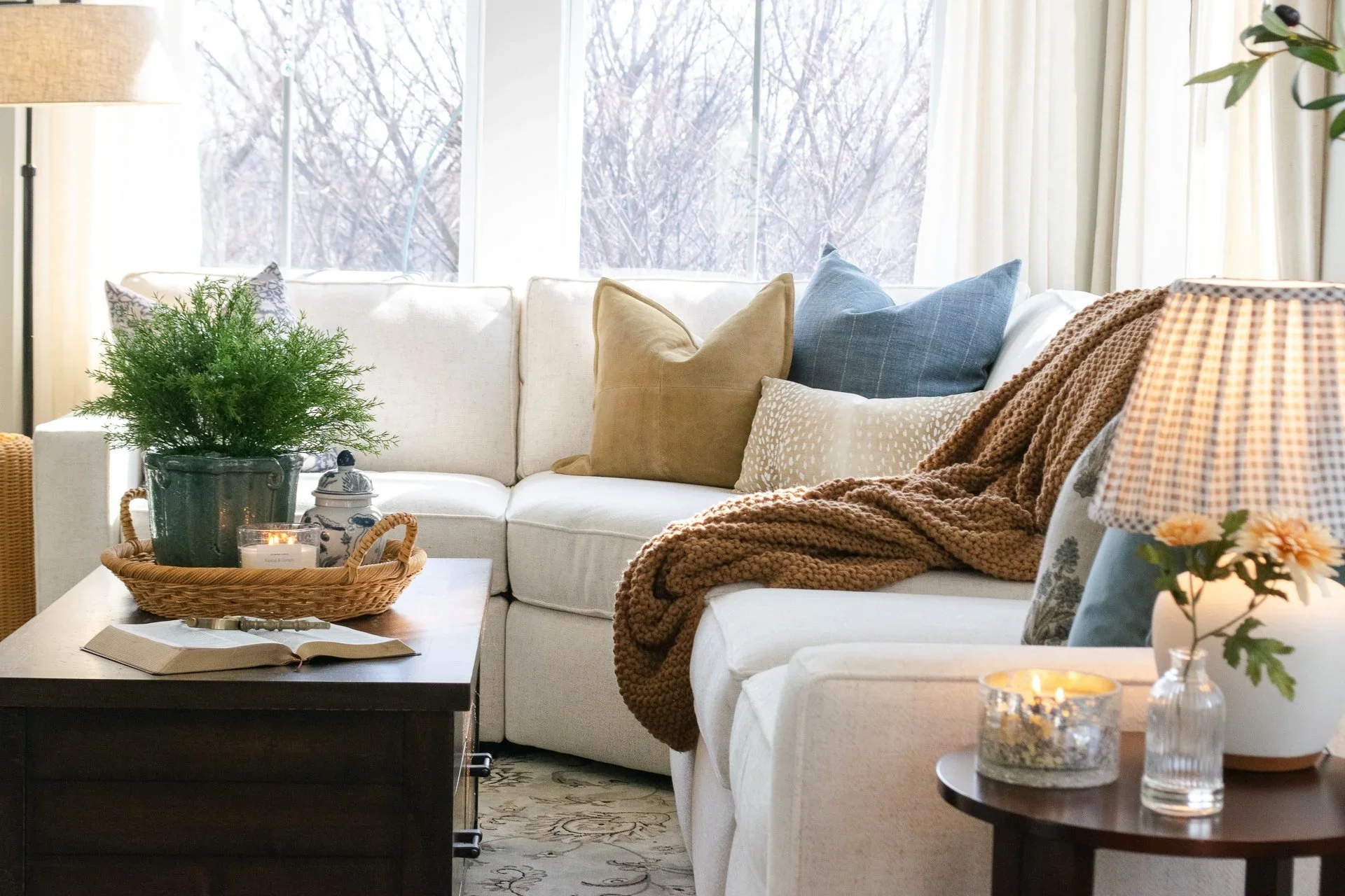 Living room with a white sectional sofa decorated with pillows and a blanket, a coffee table with a plant, candle, and book, a side table with a candle and flowers, and large windows showing trees outside.