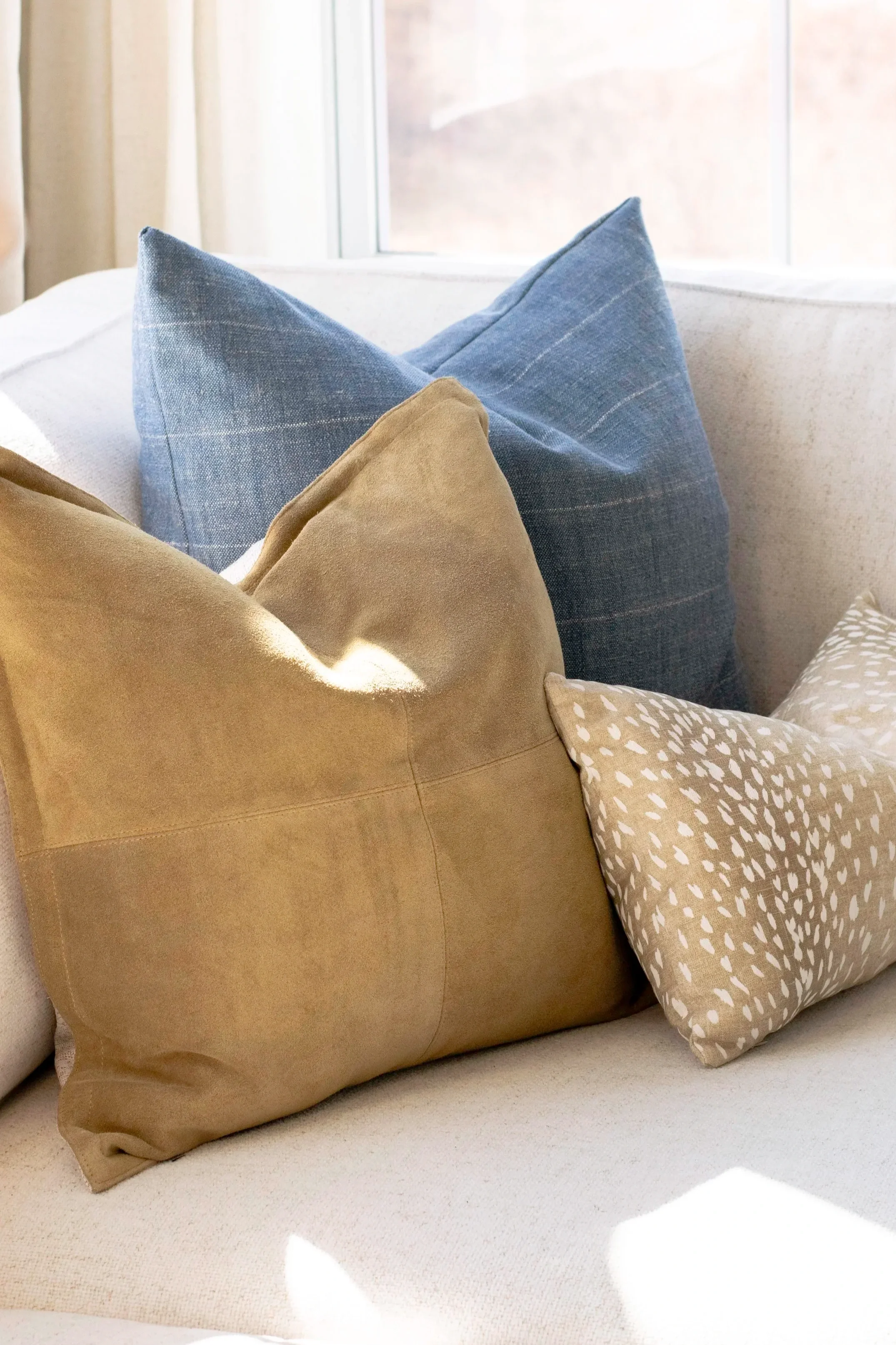 A beige sofa with a large tan cushion, a blue cushion with pinstripes, and a beige cushion with white leaf prints, near a window with curtains.