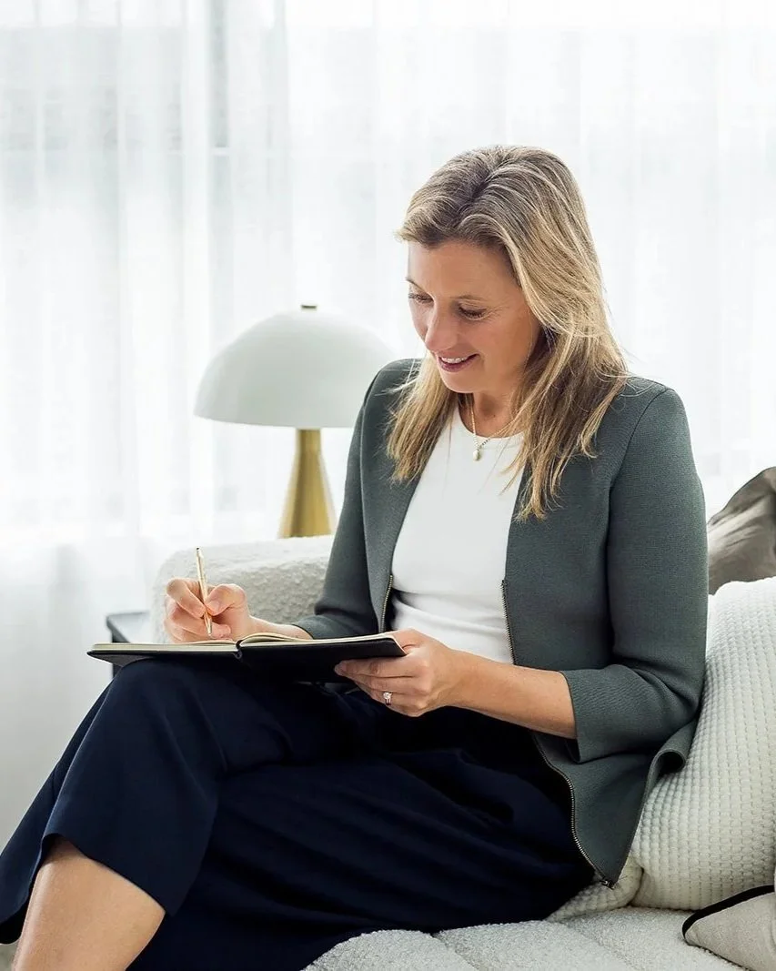 A woman sitting on a white sofa, writing in a notebook with a pen.