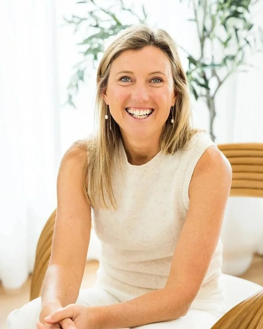 A woman with blonde hair in a sleeveless cream-colored top, smiling and sitting on a chair indoors with a background of a plant and white curtains.