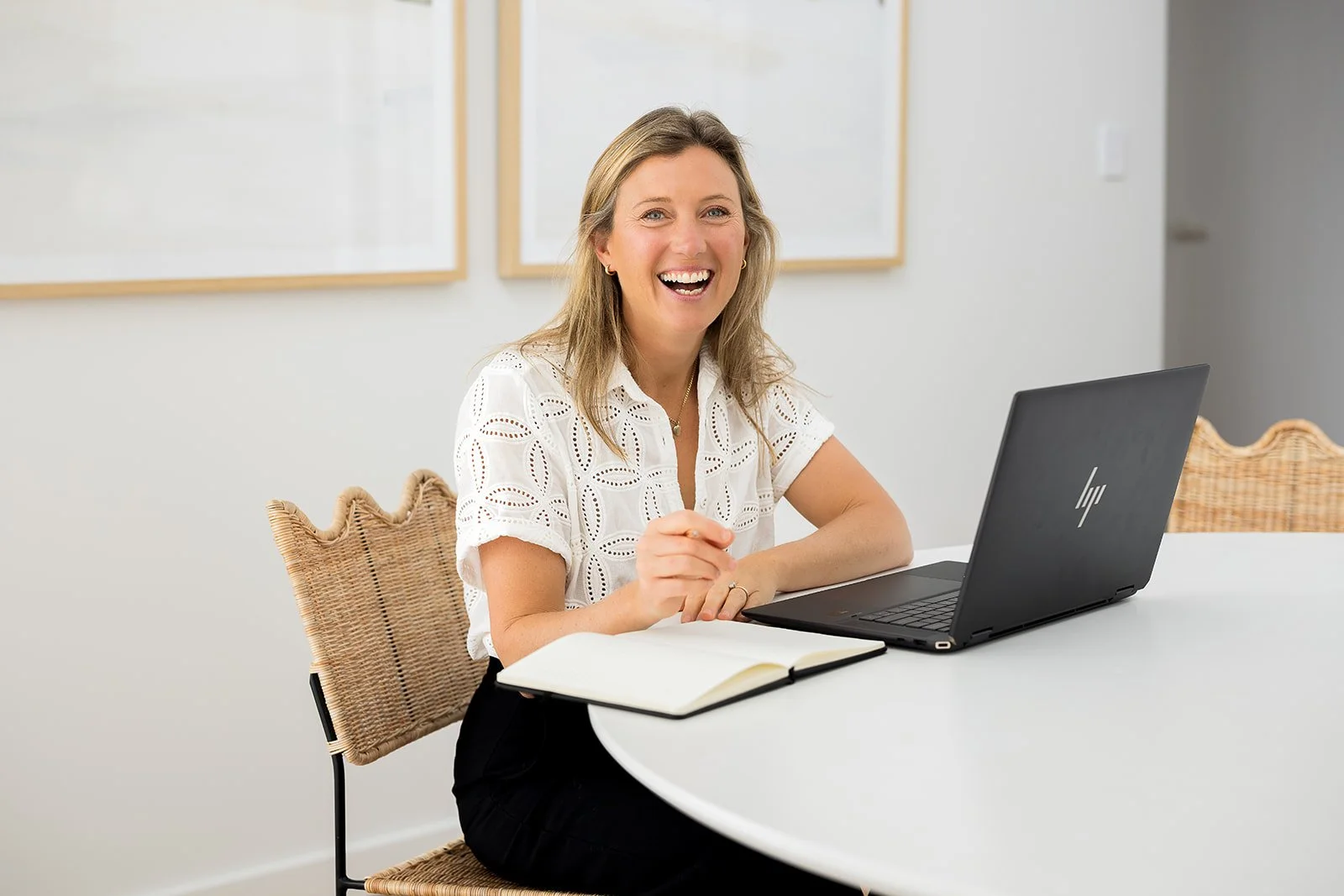 A woman sitting at a white table with a black HP laptop, smiling and looking at the camera, with a notebook open in front of her.