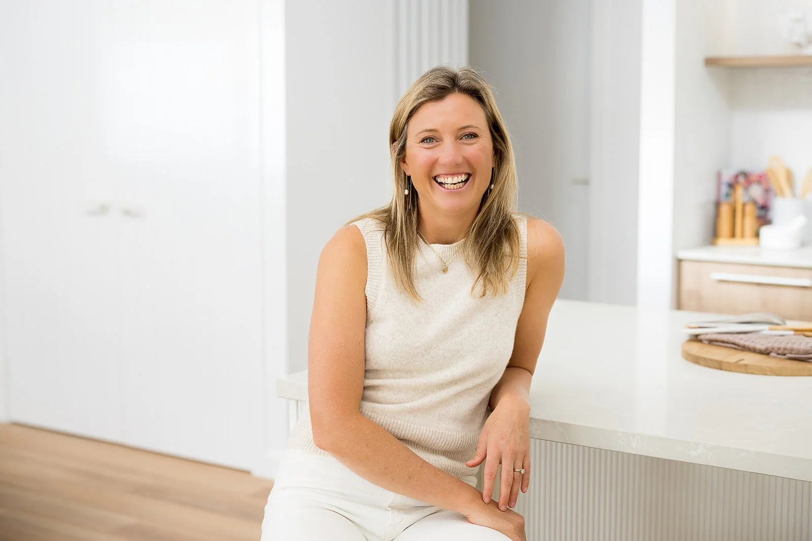 A woman with shoulder-length light brown hair and a broad smile, wearing a sleeveless cream top and white pants, sitting at a kitchen counter in a modern, bright kitchen with white and wooden accents.