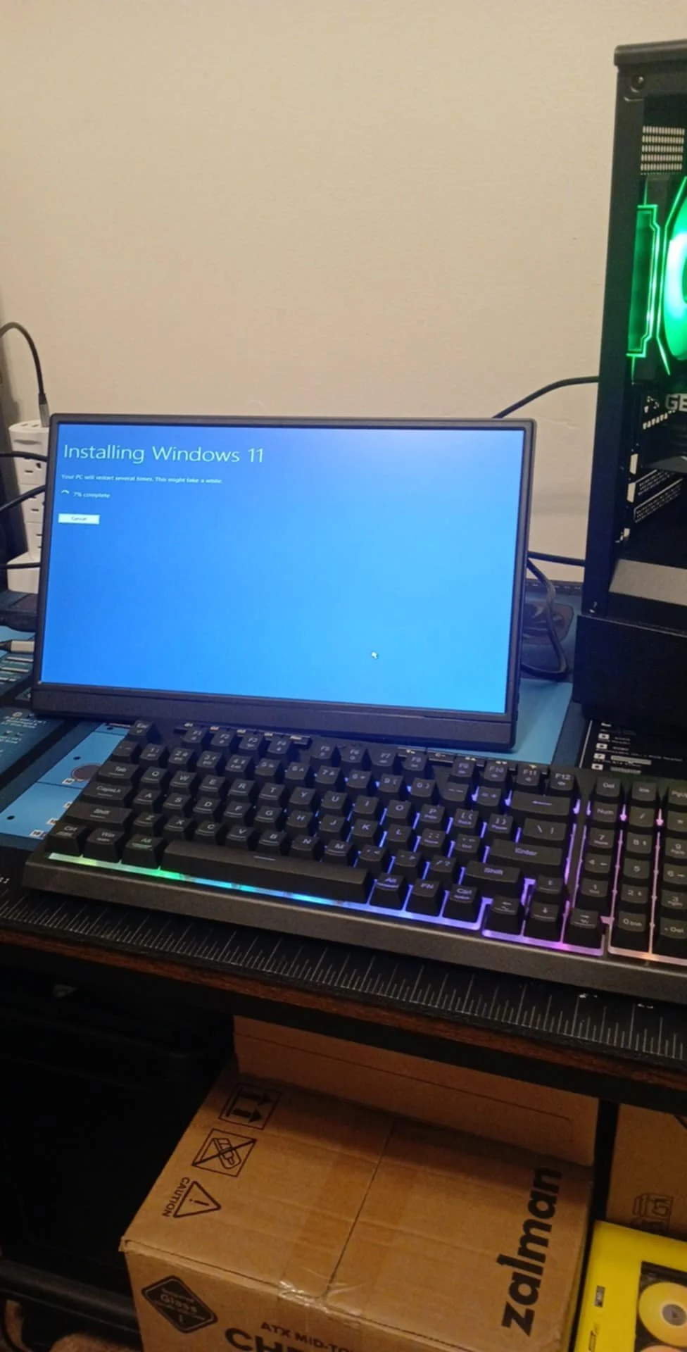A computer setup with a monitor installing Windows 11, a mechanical keyboard with backlit keys, and a gaming PC tower with green lighting.